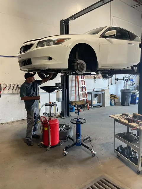 An auto mechanic inspecting the underside of a white car elevated on a hydraulic lift inside a garage or repair shop.