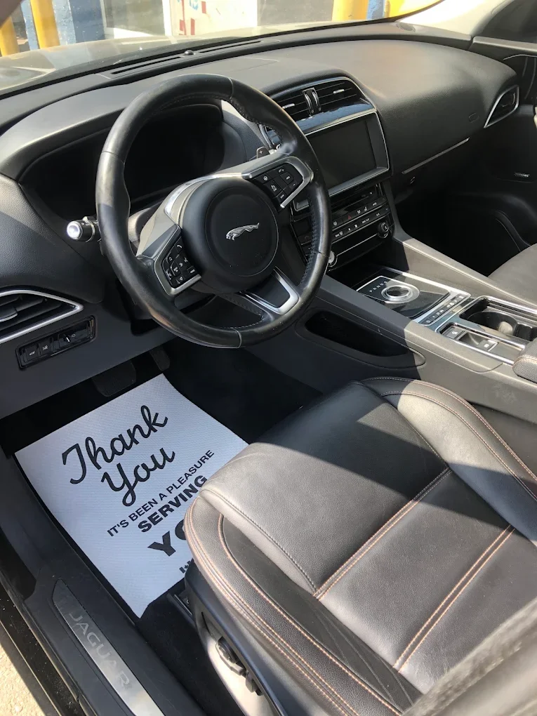 Inside view of a Jaguar vehicle's driver's seat and dashboard, featuring a leather seat with orange stitching, a steering wheel with a Jaguar logo, a digital display, and a floor mat saying 'Thank You, It’s Been a Pleasure Serving You'.