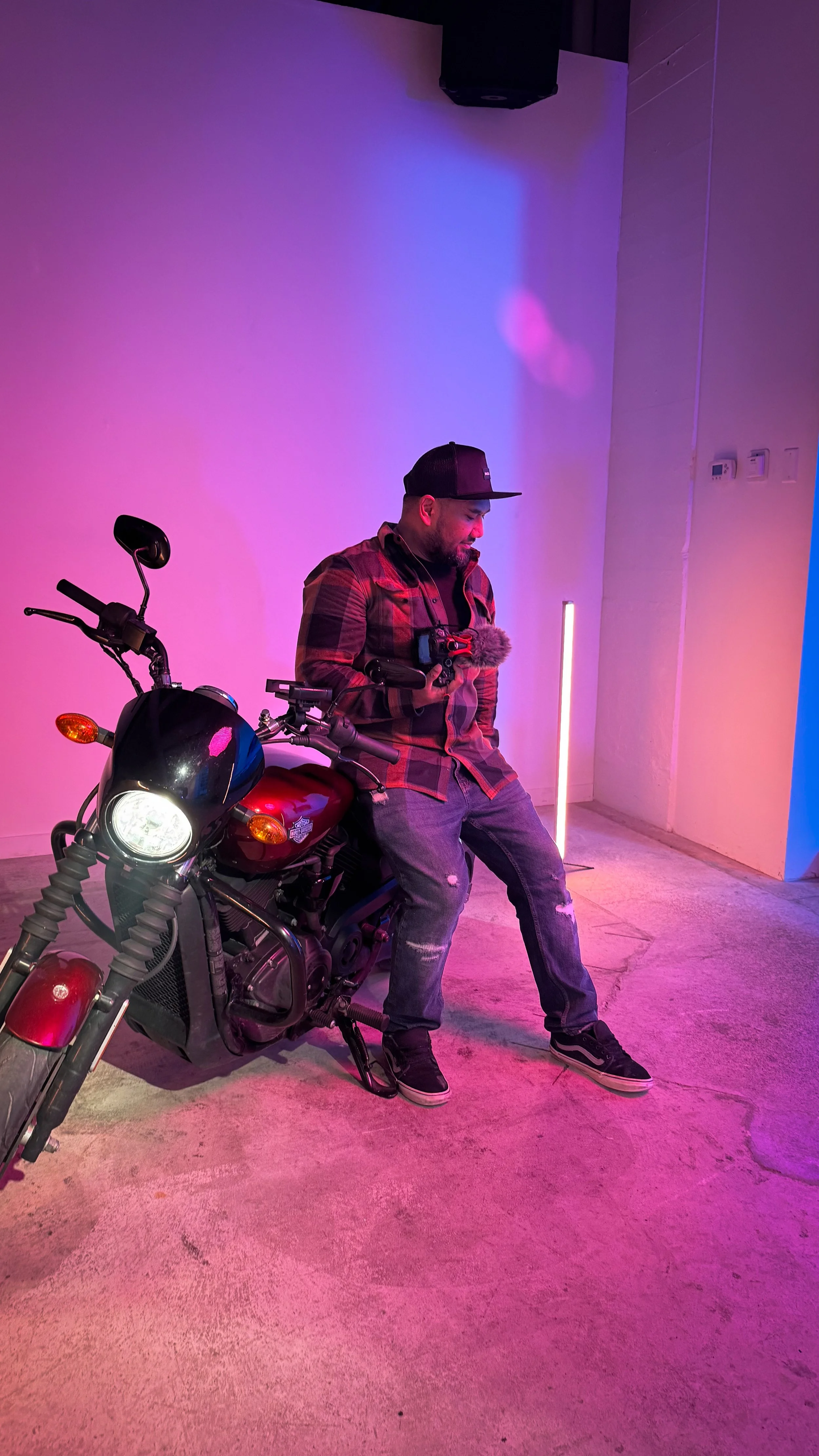 A man sitting on a red motorcycle with black details in a room with purple lighting. He is wearing a black cap, a red and black plaid shirt, gray ripped jeans, and black sneakers. The room has a plain wall, a vertical light, and some electrical outlets.