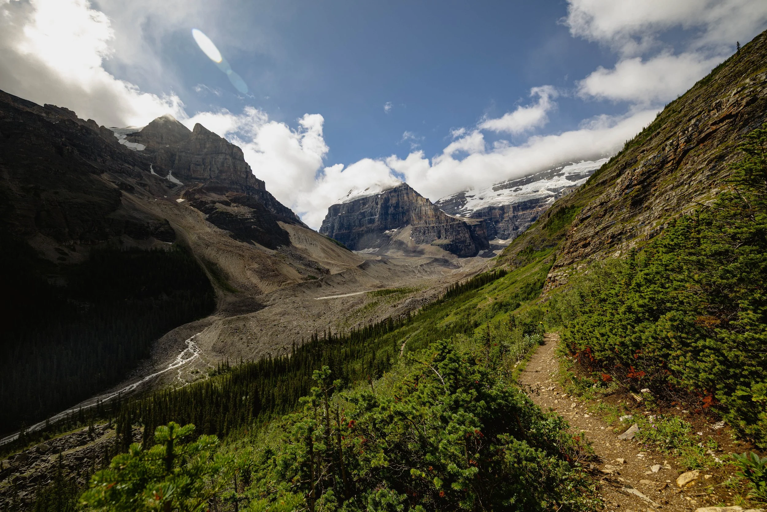A mountain landscape with rugged peaks partially covered in snow, green trees in the foreground on a clear day with some clouds in the sky.