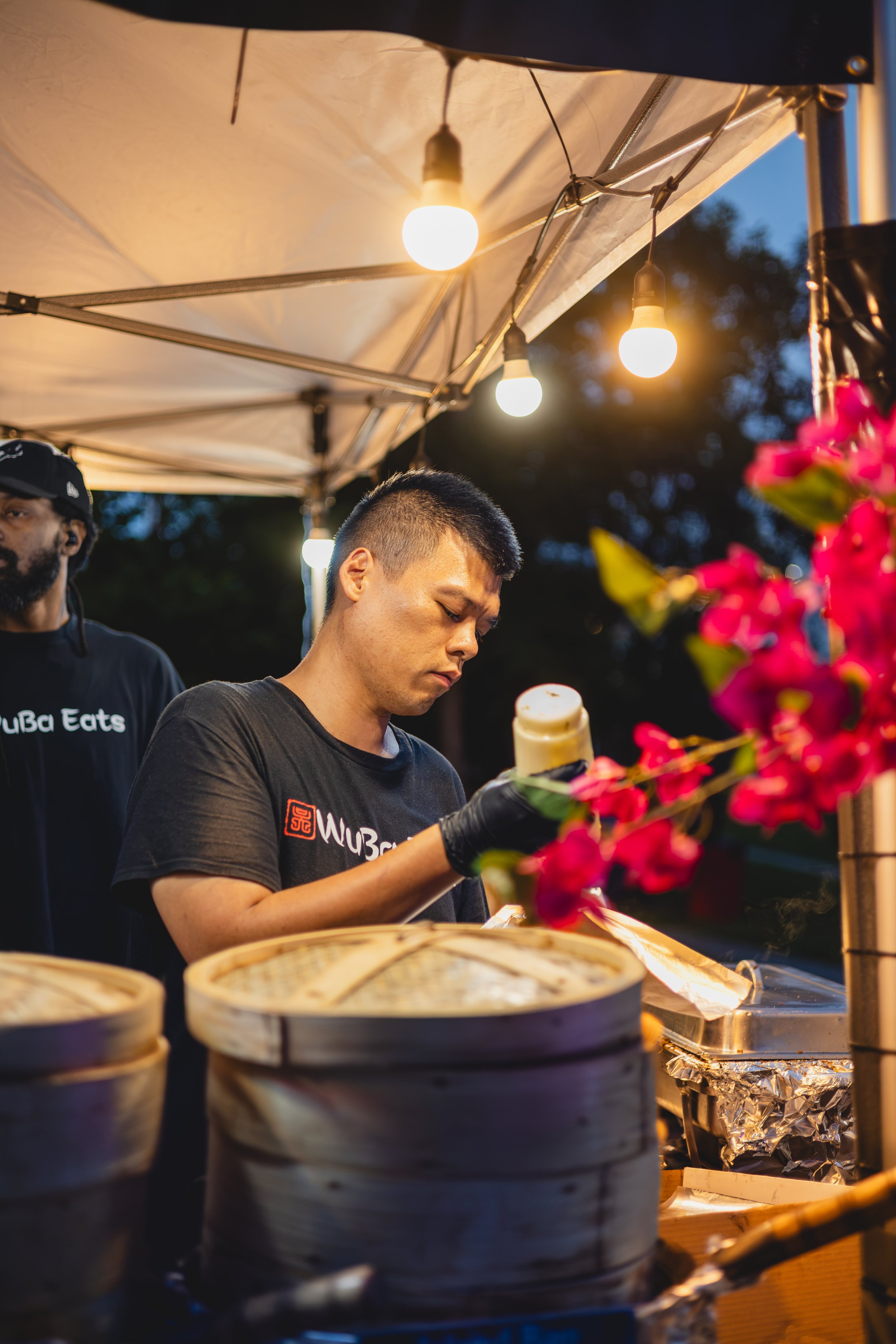 A man preparing food under a canopy with string lights hanging above, with bamboo steamers in the foreground and pink flowers on the side, in an outdoor setting during evening.