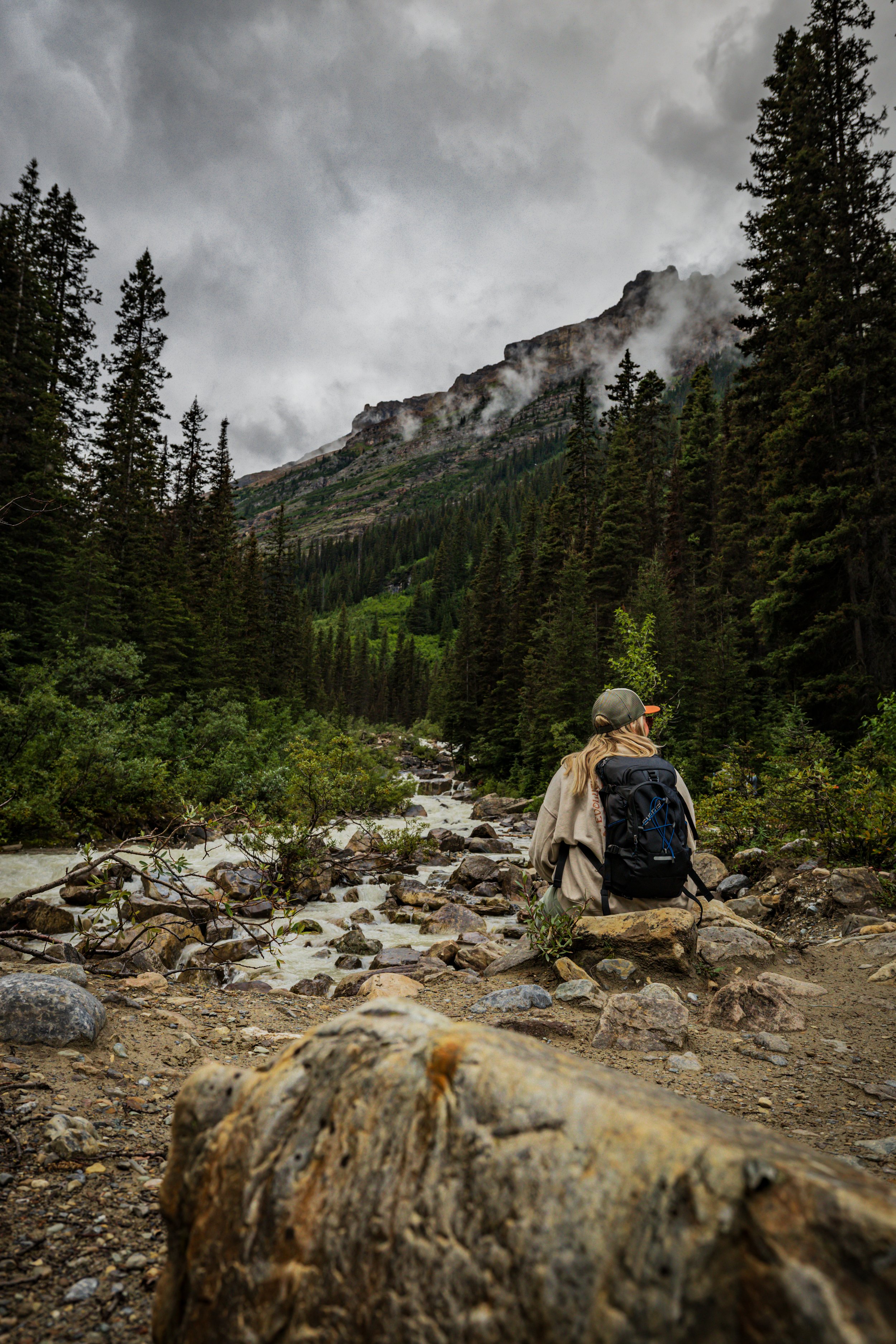 A person sitting on rocks near a river in a dense forest with mountains and cloudy sky in the background.