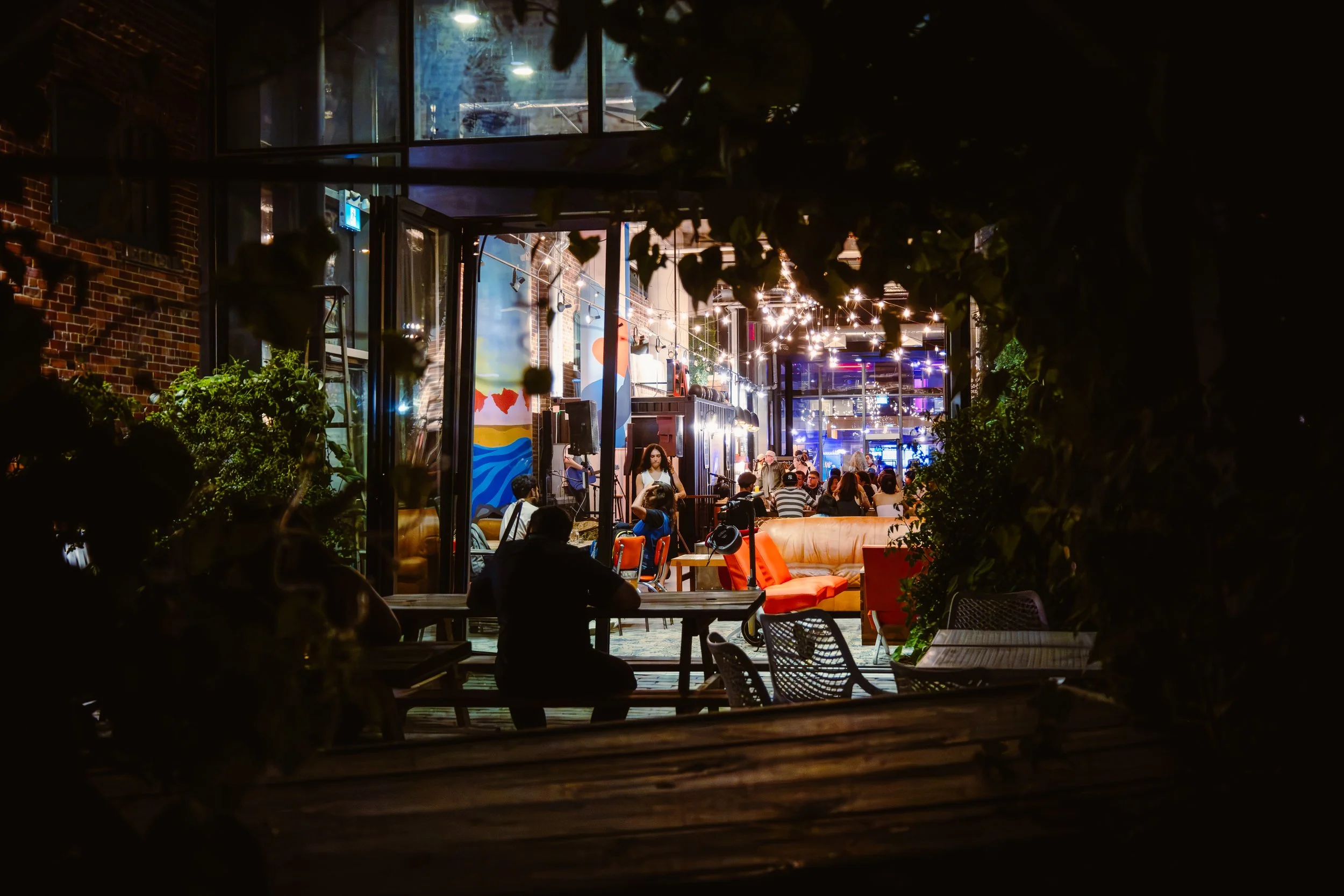 Night view of a busy indoor restaurant or café with people seated at tables, colorful wall art, and string lights, framed by dark green foliage in the foreground.