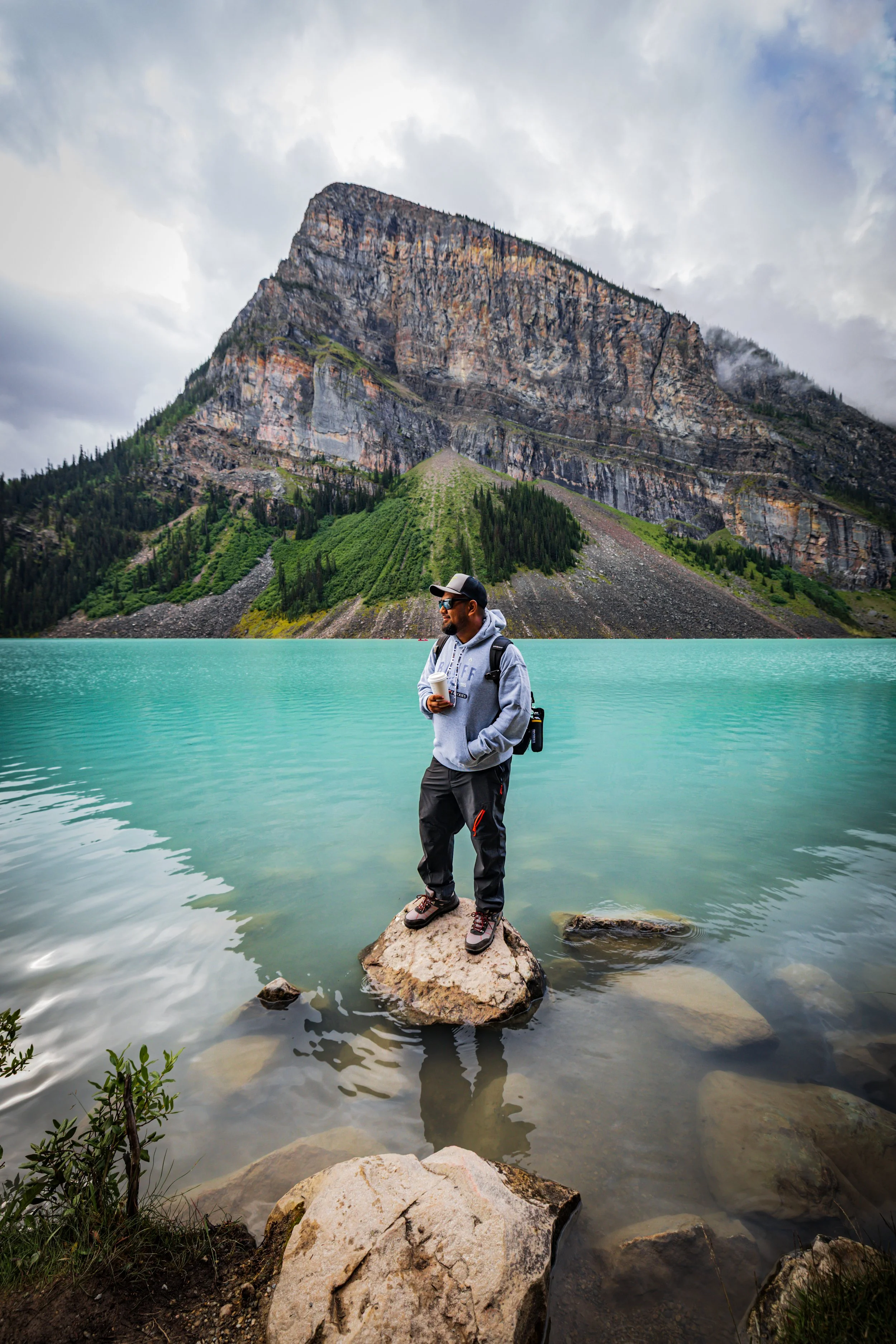 Man standing on rock in turquoise lake with mountain background and cloudy sky, holding coffee cup and wearing sunglasses, hoodie, and hiking boots.