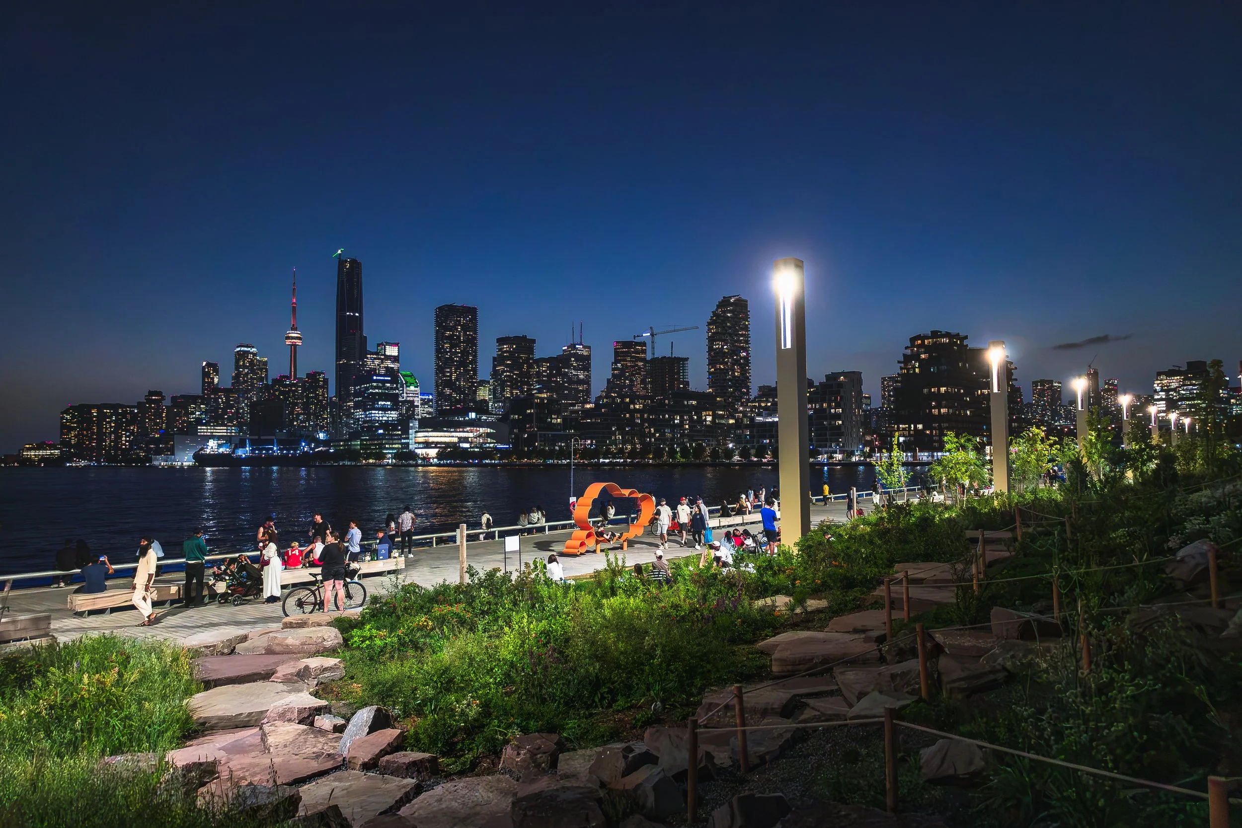 Nighttime city skyline with illuminated skyscrapers along a waterfront. People relax and walk in a park area with trees, rocks, and walking paths in the foreground.