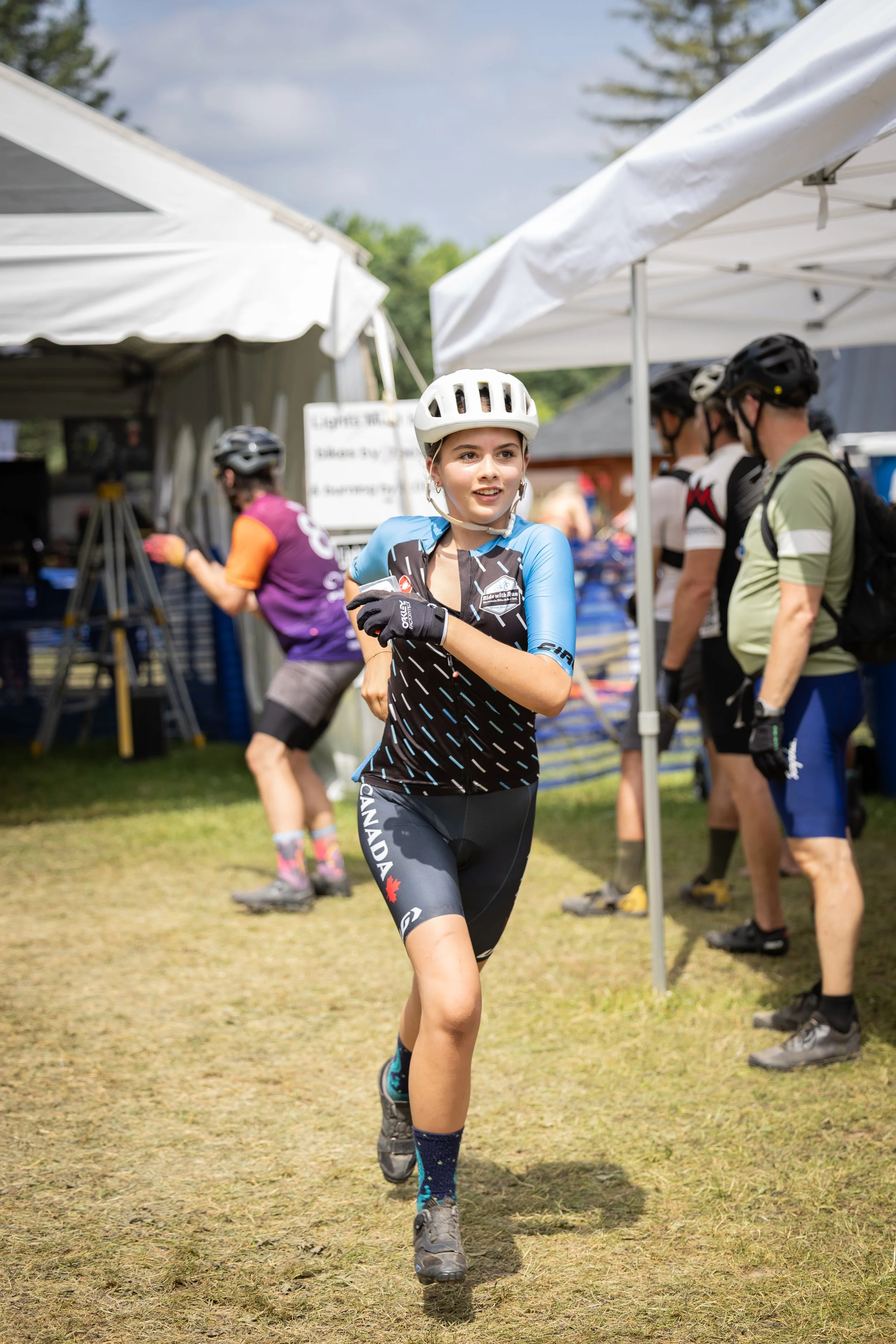 Young woman in cycling gear and helmet running on grass at an outdoor event, with people and tents in the background.