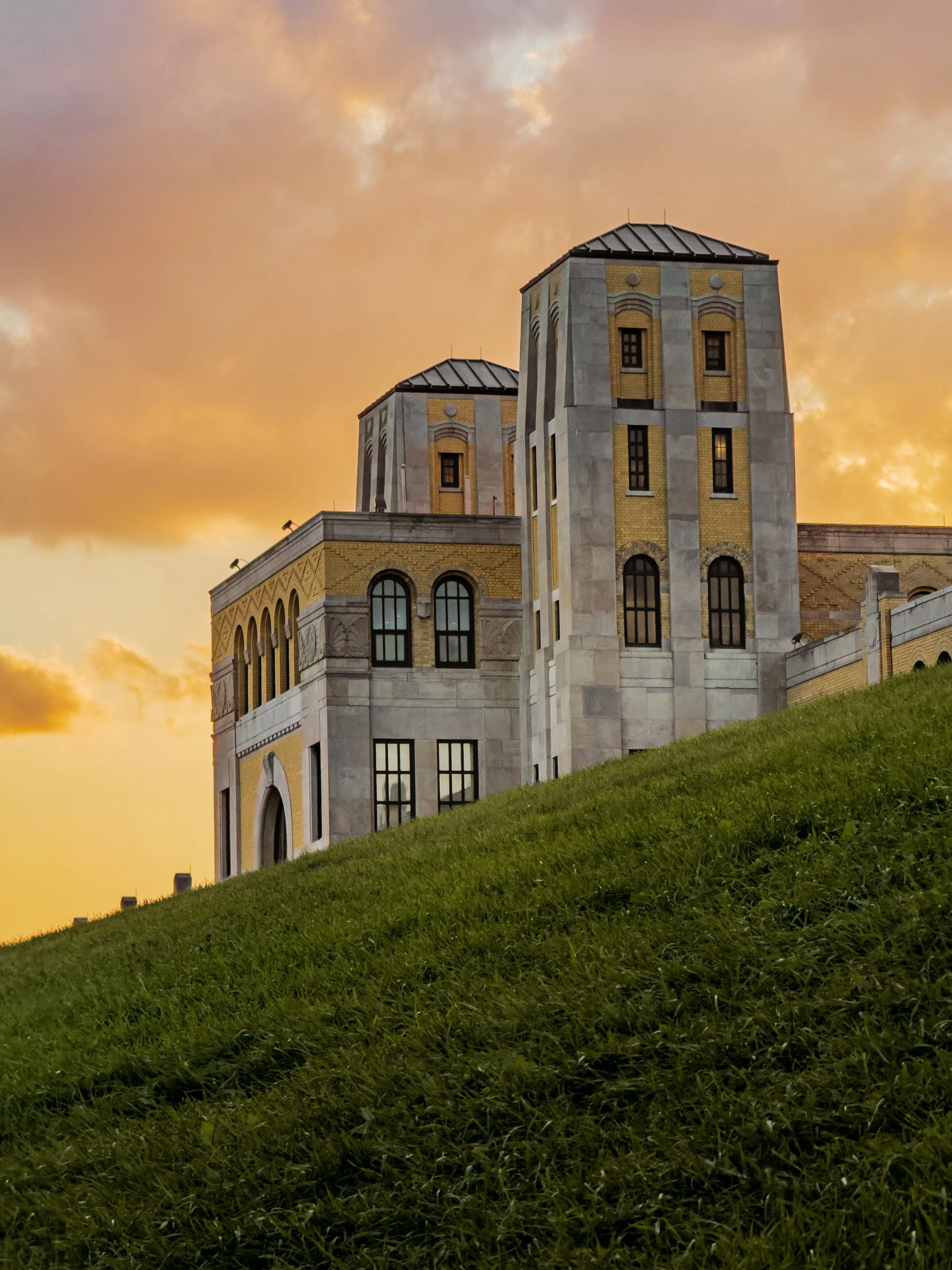 A large castle-like building with tall towers and arched windows, situated on a grassy hillside during sunset with a partly cloudy orange sky.