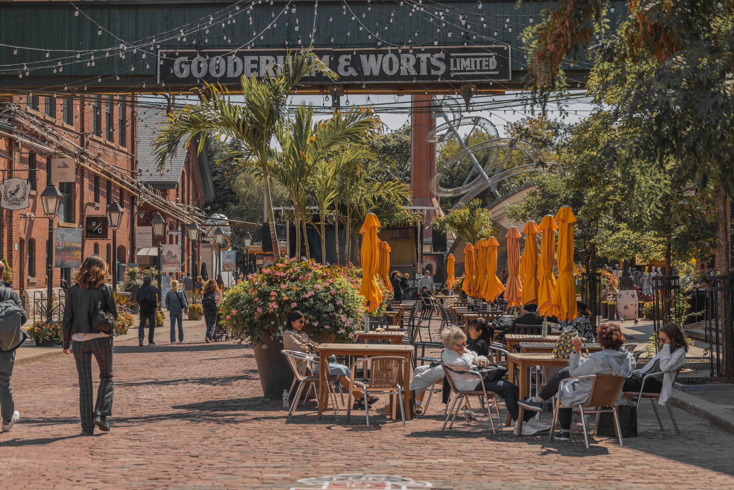 People sitting and walking in an outdoor dining area with yellow umbrellas and large flower pots, on a sunny day in a city street lined with brick buildings and trees, with a sign that reads 'Gooder & Worts Limited' and string lights overhead.