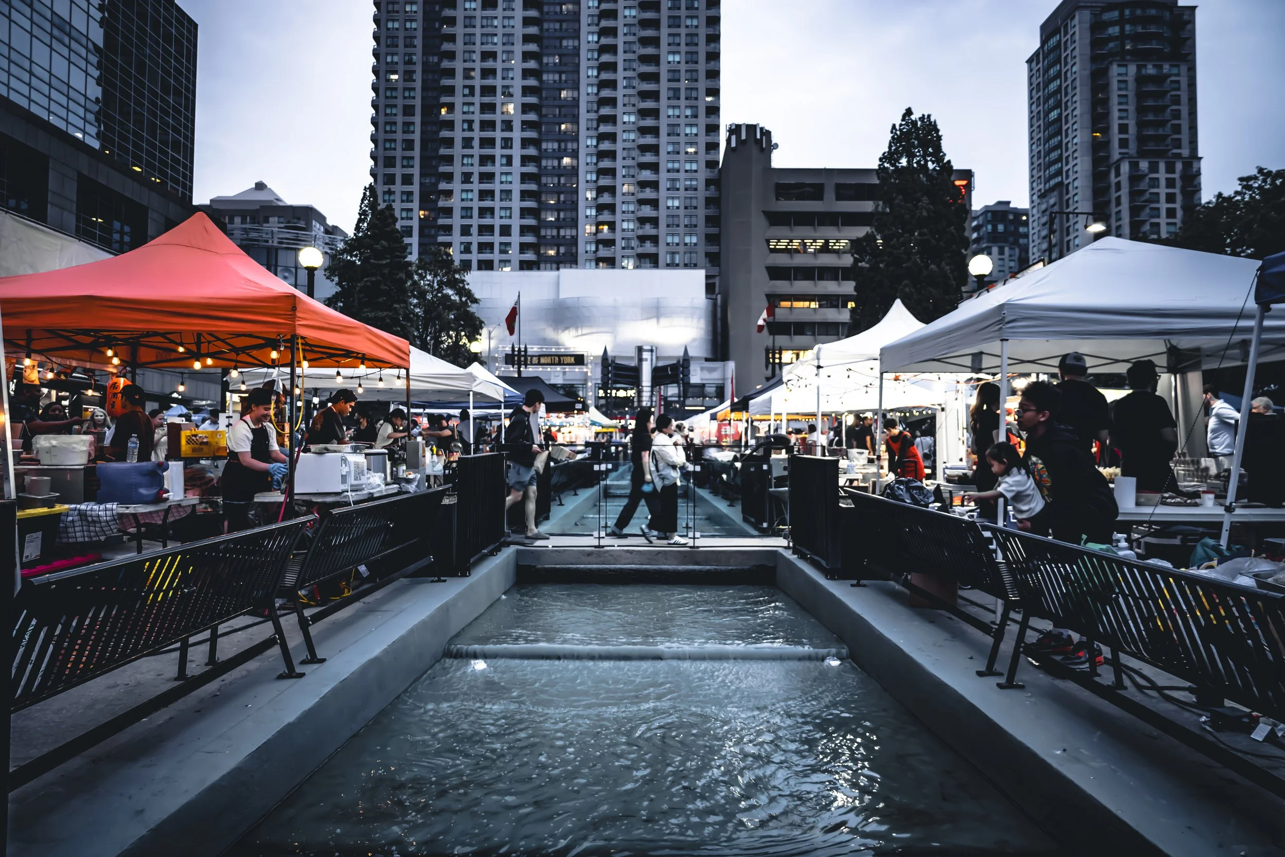 Outdoor marketplace with white and orange tents, people shopping and selling, tall buildings in the background, and water feature in the foreground, during dusk.