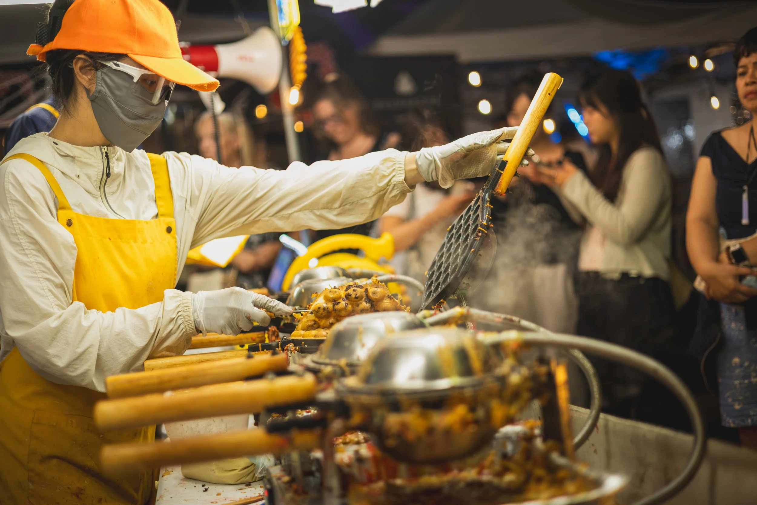 A person wearing a yellow apron, gray face mask, and orange cap serving food at a buffet station with multiple trays of cooked food, surrounded by others in a dimly lit setting.