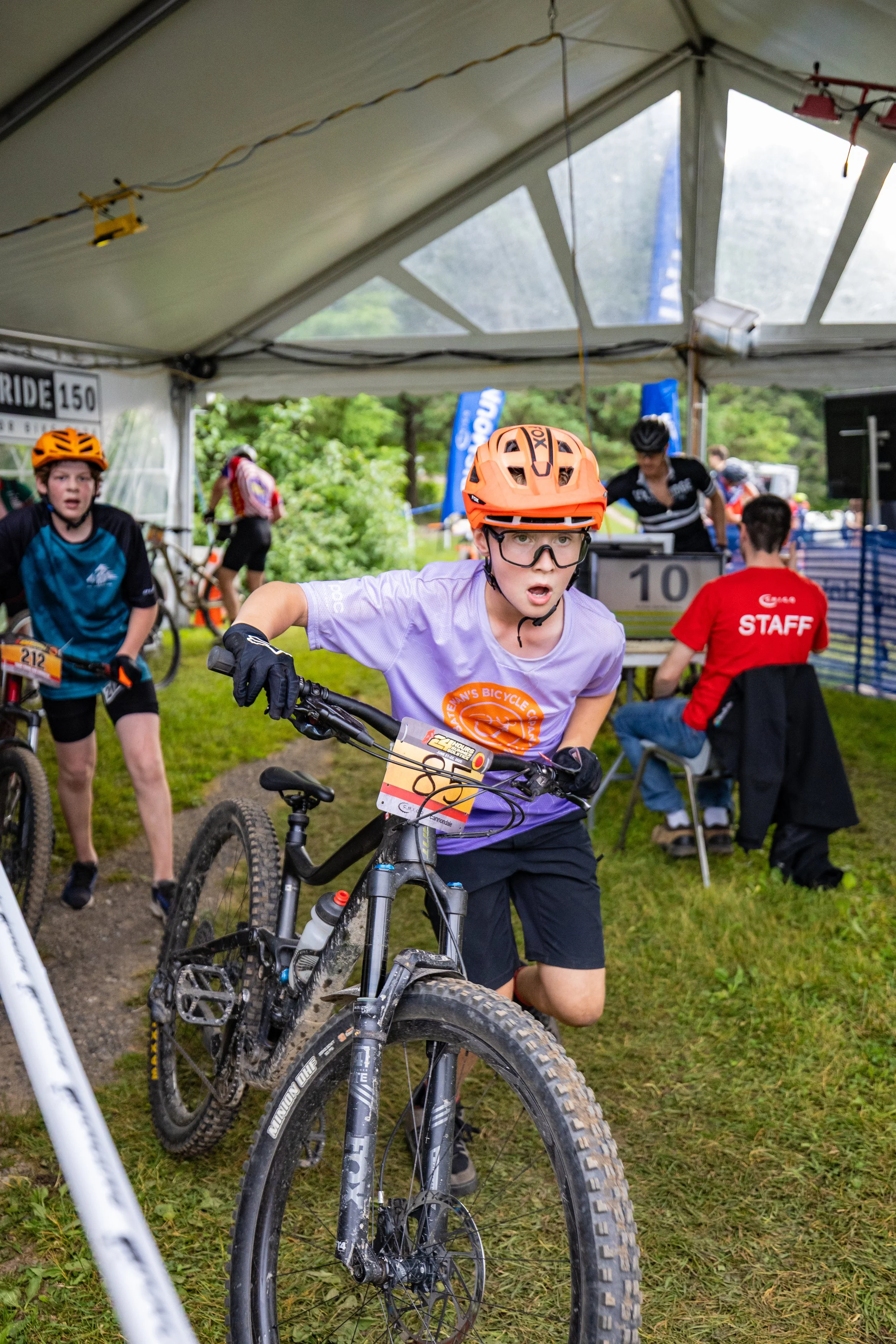 Young boy with a mountain bike, wearing a orange helmet and glasses, participating in a biking event under a tent with other riders and event staff visible in the background.