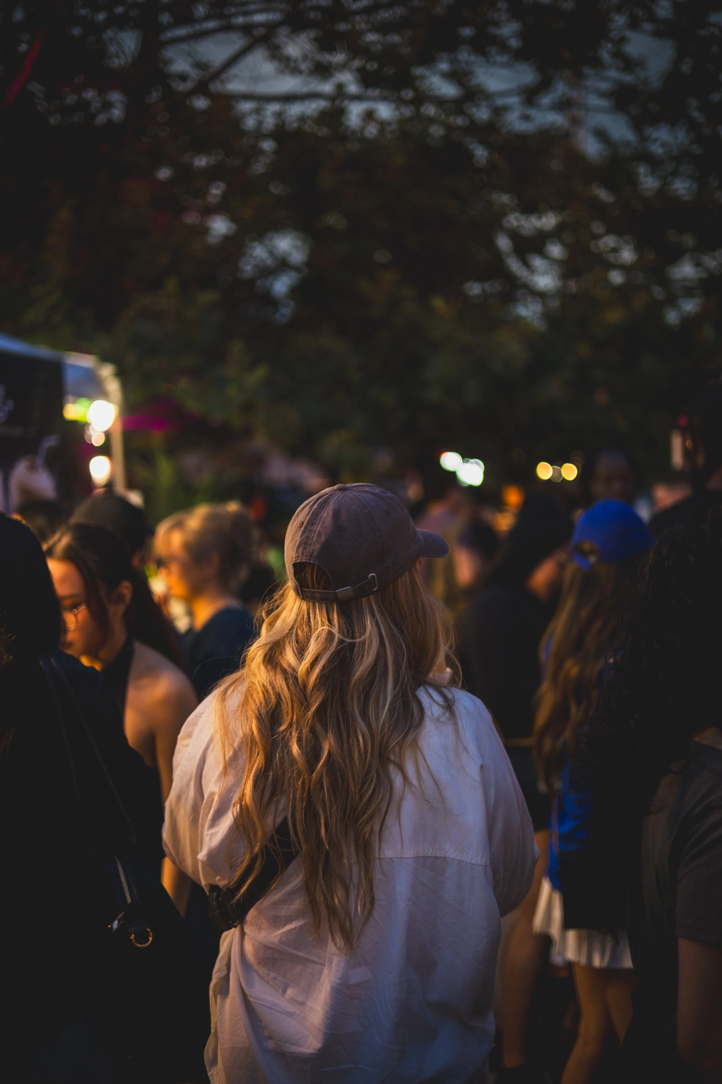 People gathered outdoors during the evening, with some individuals wearing hats and jackets, under a large tree with scattered lights and a darkening sky.