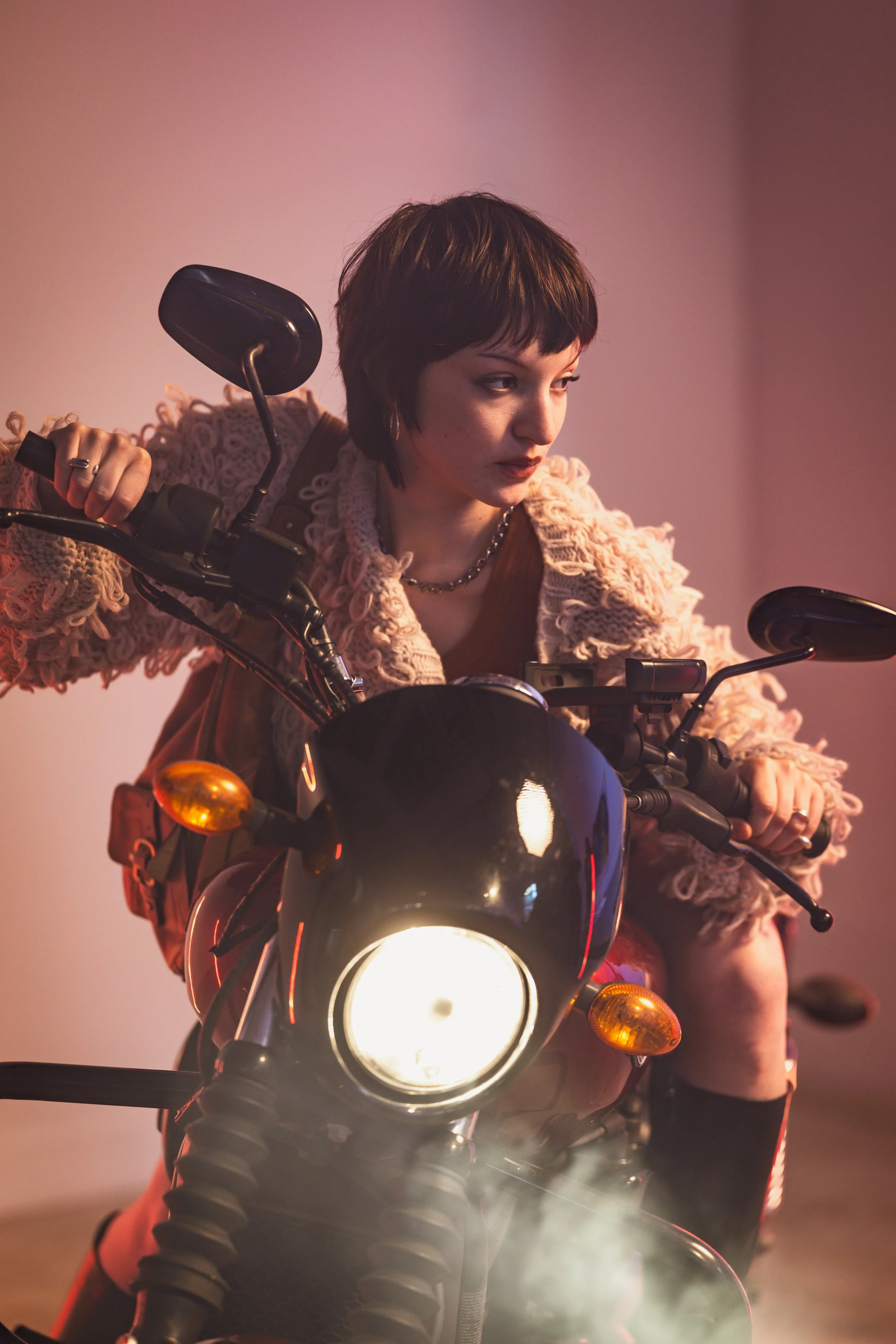 A woman with short dark hair, wearing a chunky pink sweater, sits on a motorcycle indoors with a plain light-colored wall in the background. She is looking to her right, with her hands on the motorcycle handles, and the headlight is turned on.