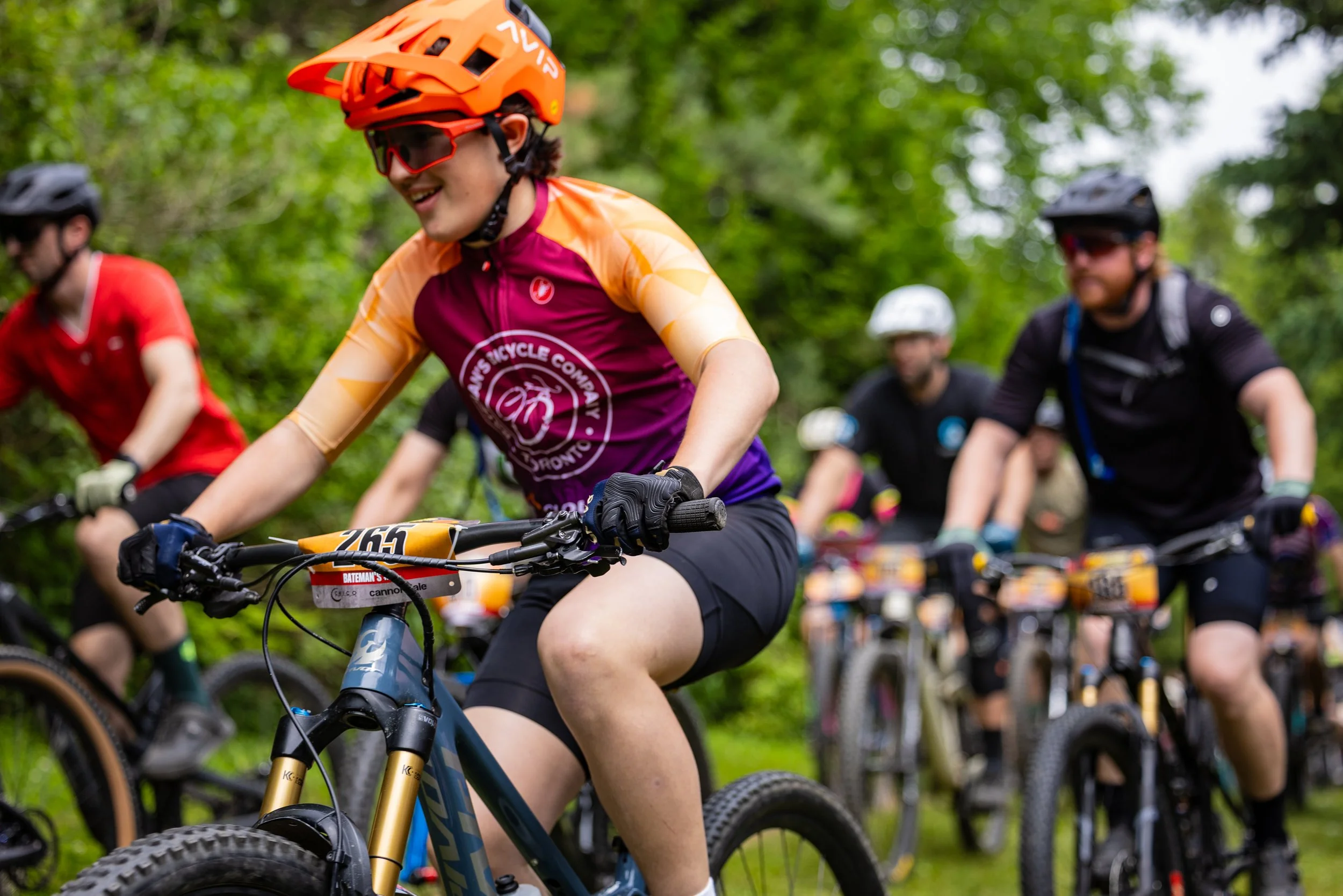 A group of cyclists riding mountain bikes through a wooded area during a race or event. The focus is on a smiling woman wearing an orange helmet, red sunglasses, and a maroon and orange cycling jersey.