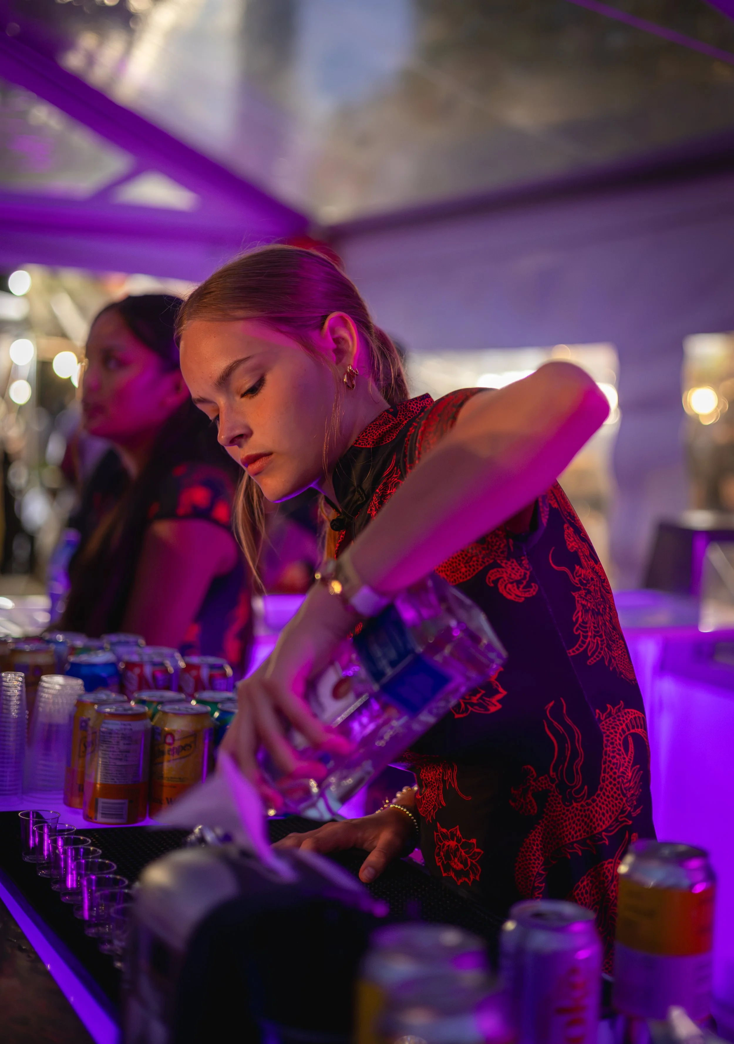 A woman with blonde hair and earrings is pouring a clear drink into a plastic cup at a bar with purple lighting. Other women are in the background, and cans of soda and plastic cups are on the bar.
