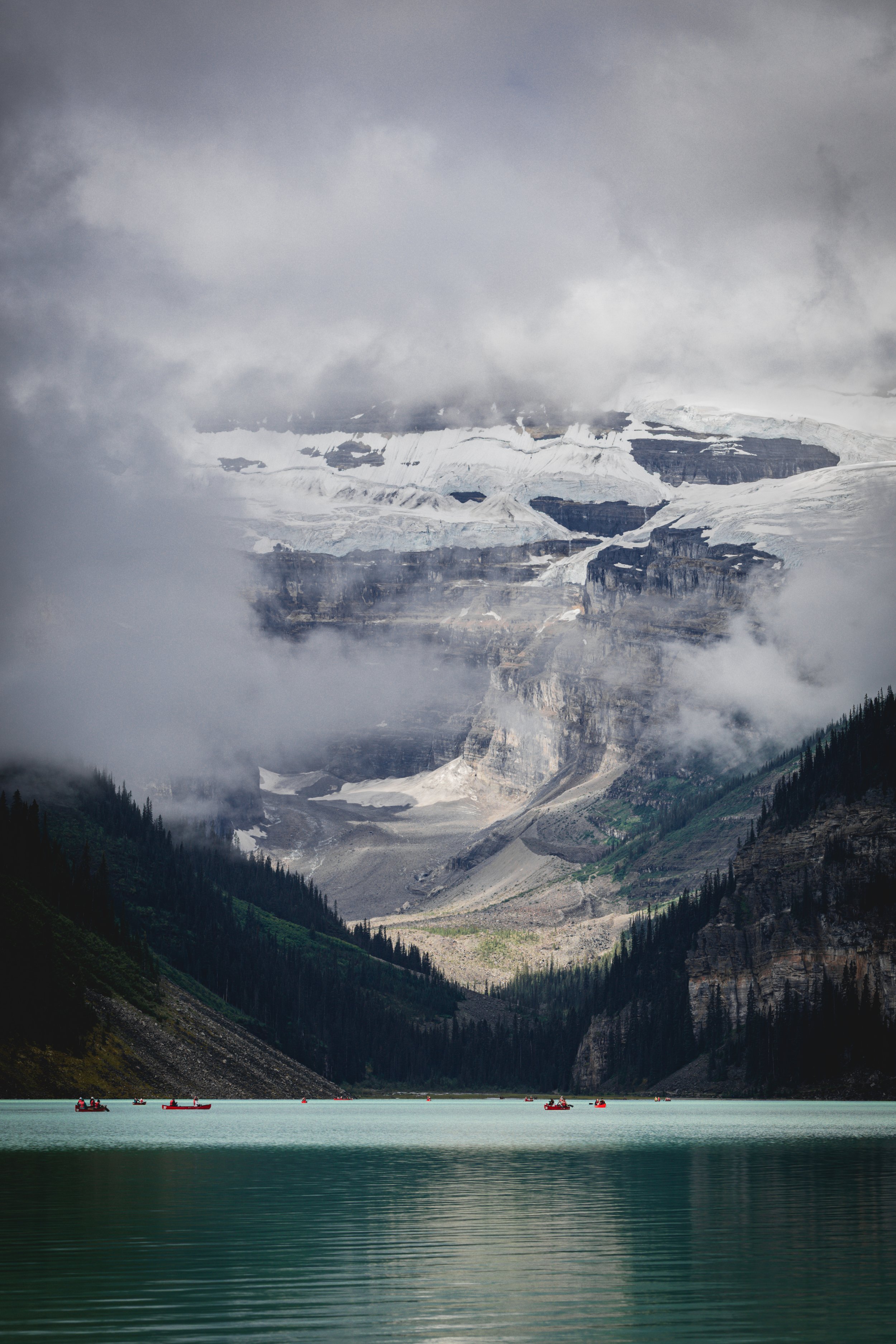A body of water with a mountain range in the background, covered in snow and ice, partly obscured by clouds and mist, with lush green trees on the shoreline.