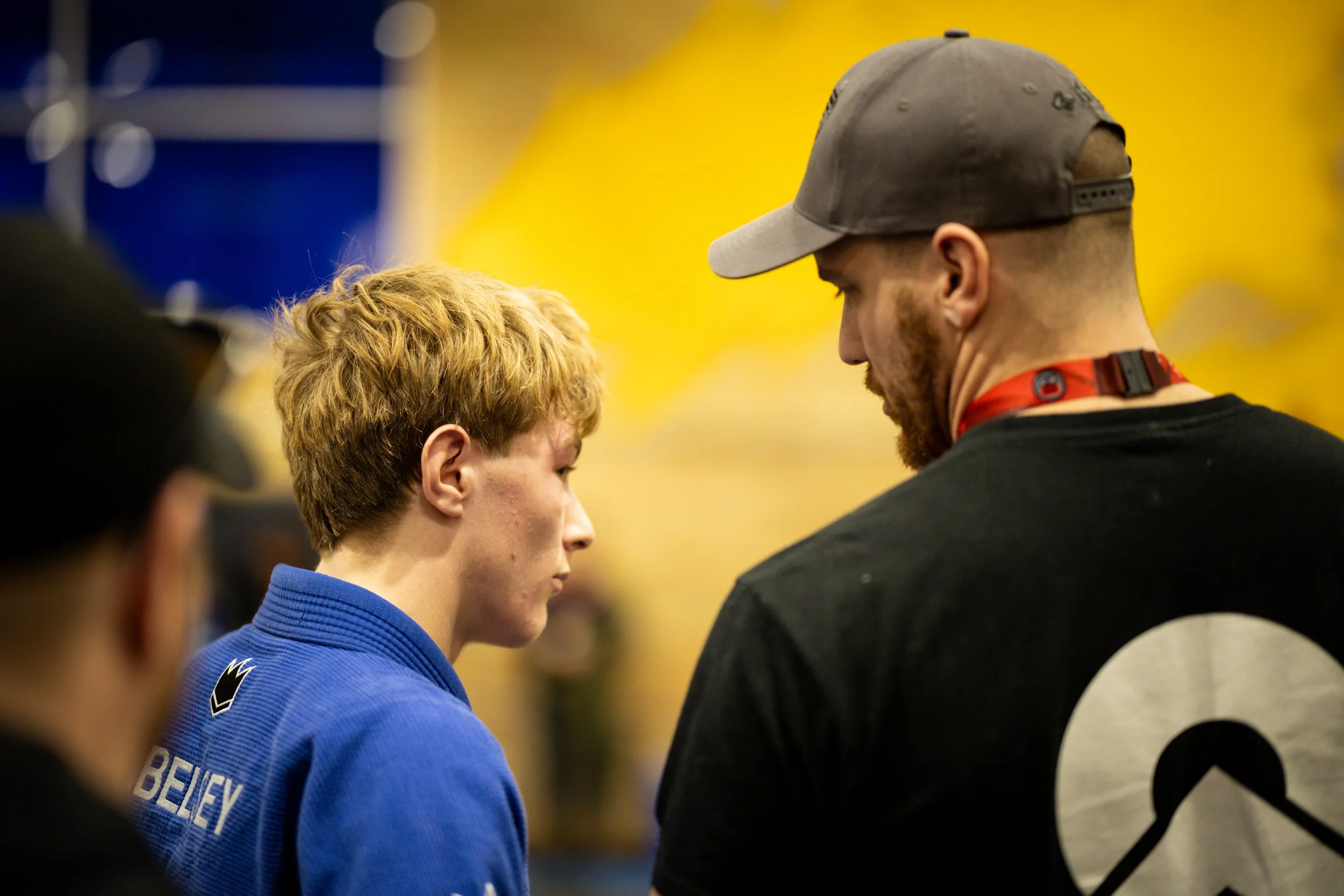 A young man in a blue martial arts uniform standing in profile, engaged in a conversation with another man in a black and white shirt and gray baseball cap, against a yellow background.