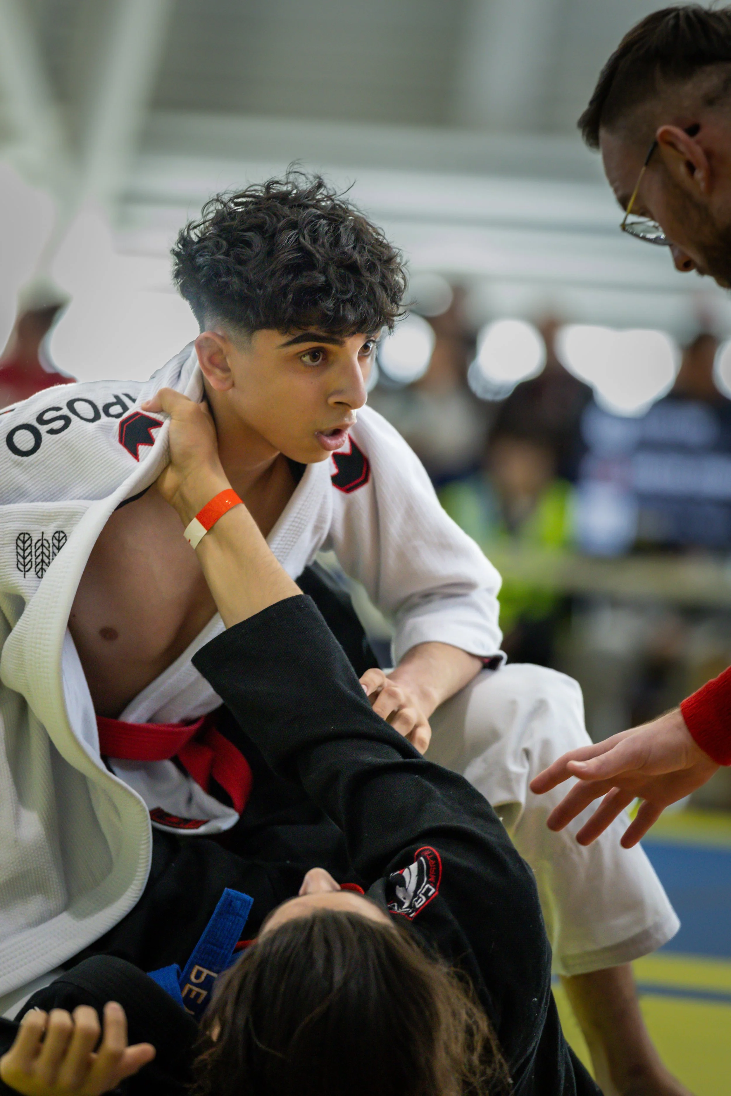Two young men dressed in jiu-jitsu gis, one with curly dark hair and light skin, and the other with short dark hair and glasses, sitting and talking in an indoor martial arts training facility.