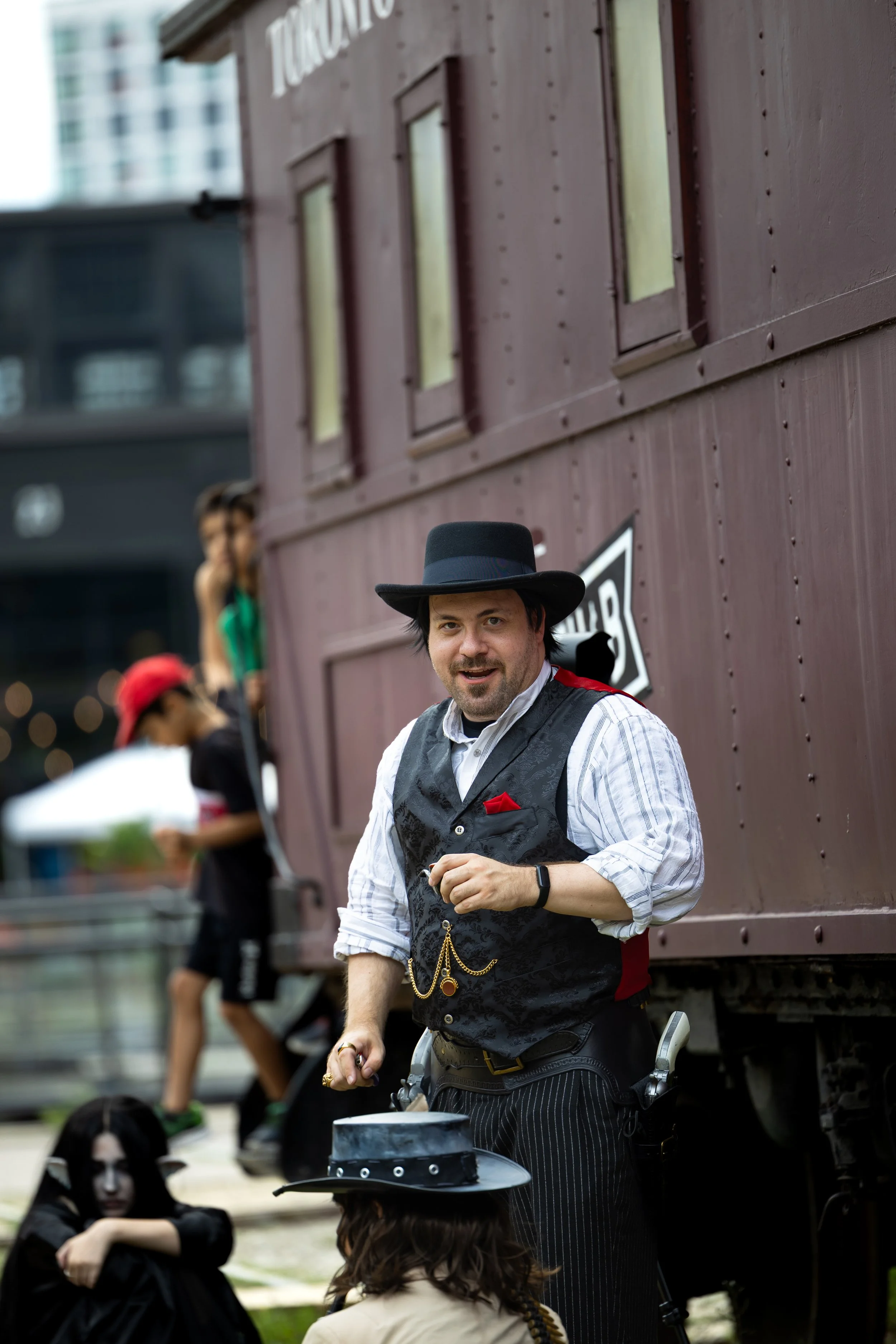 A man dressed in vintage attire, wearing a black hat and a vest with a pocket watch chain, stands in front of a brown train car. There are blurred people in the background, including a woman with a red hat and a person with long dark hair. The settin