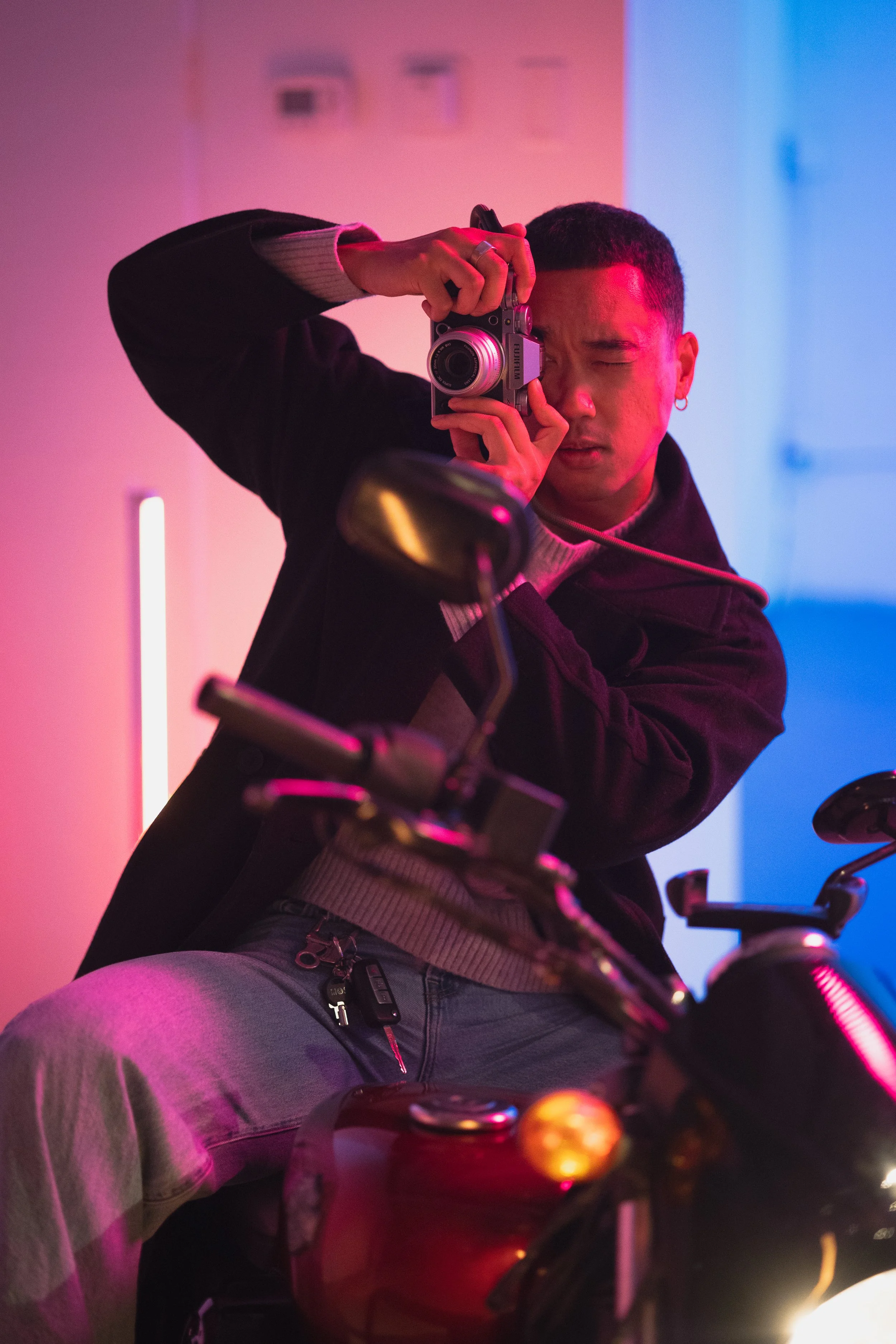 A person with short hair and a hoop earring, taking a photo with a camera in a room with pink and blue lighting, sitting on a motorcycle.