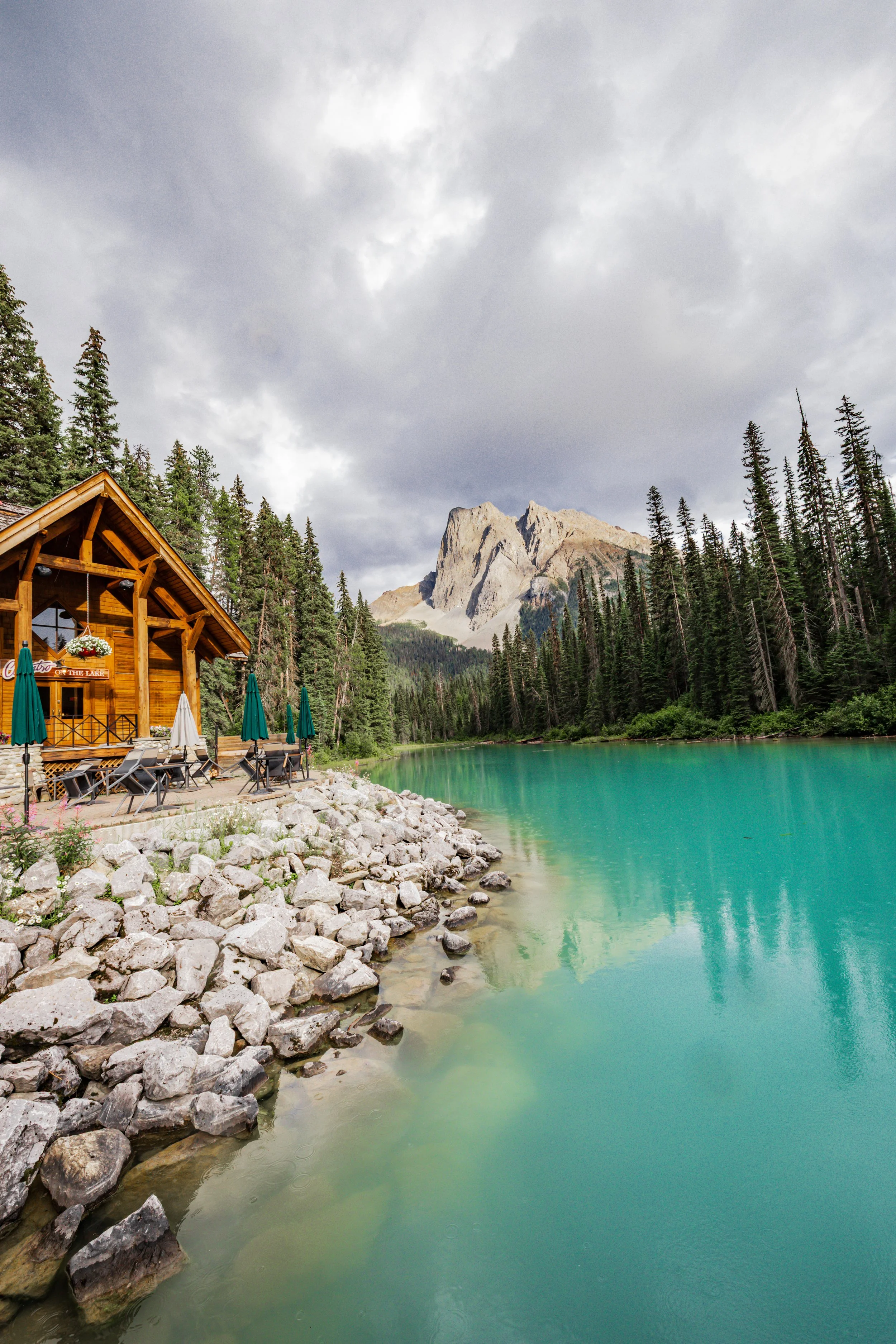 A wooden cabin with outdoor seating and umbrellas by a turquoise lake, surrounded by pine trees and mountains under cloudy sky.