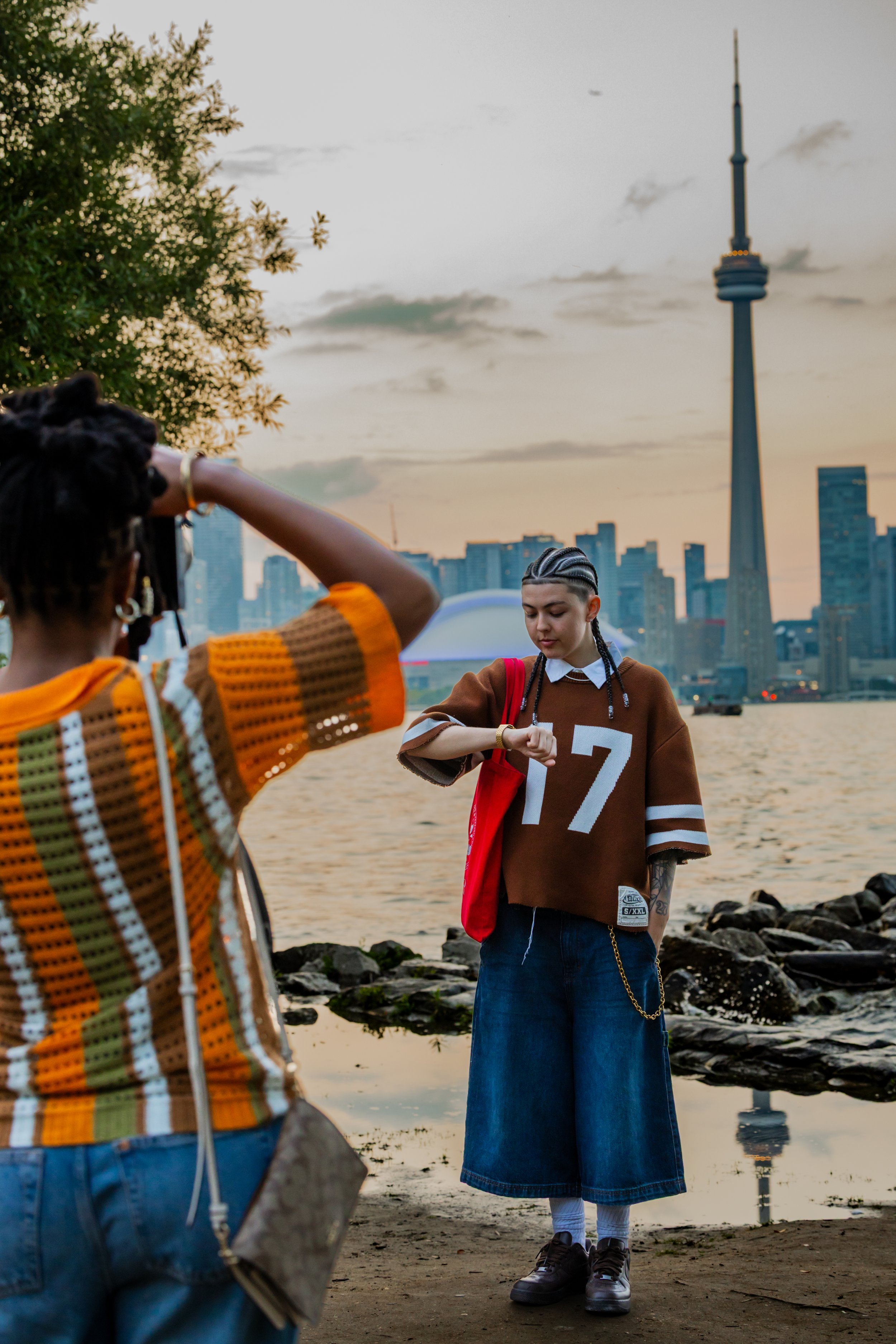 Young woman with braided hair wearing a large brown T-shirt with the number 7, blue denim skirt, and sneakers stands near water, with the Toronto skyline and CN Tower in the background during sunset. Another person, partially visible, is taking her p