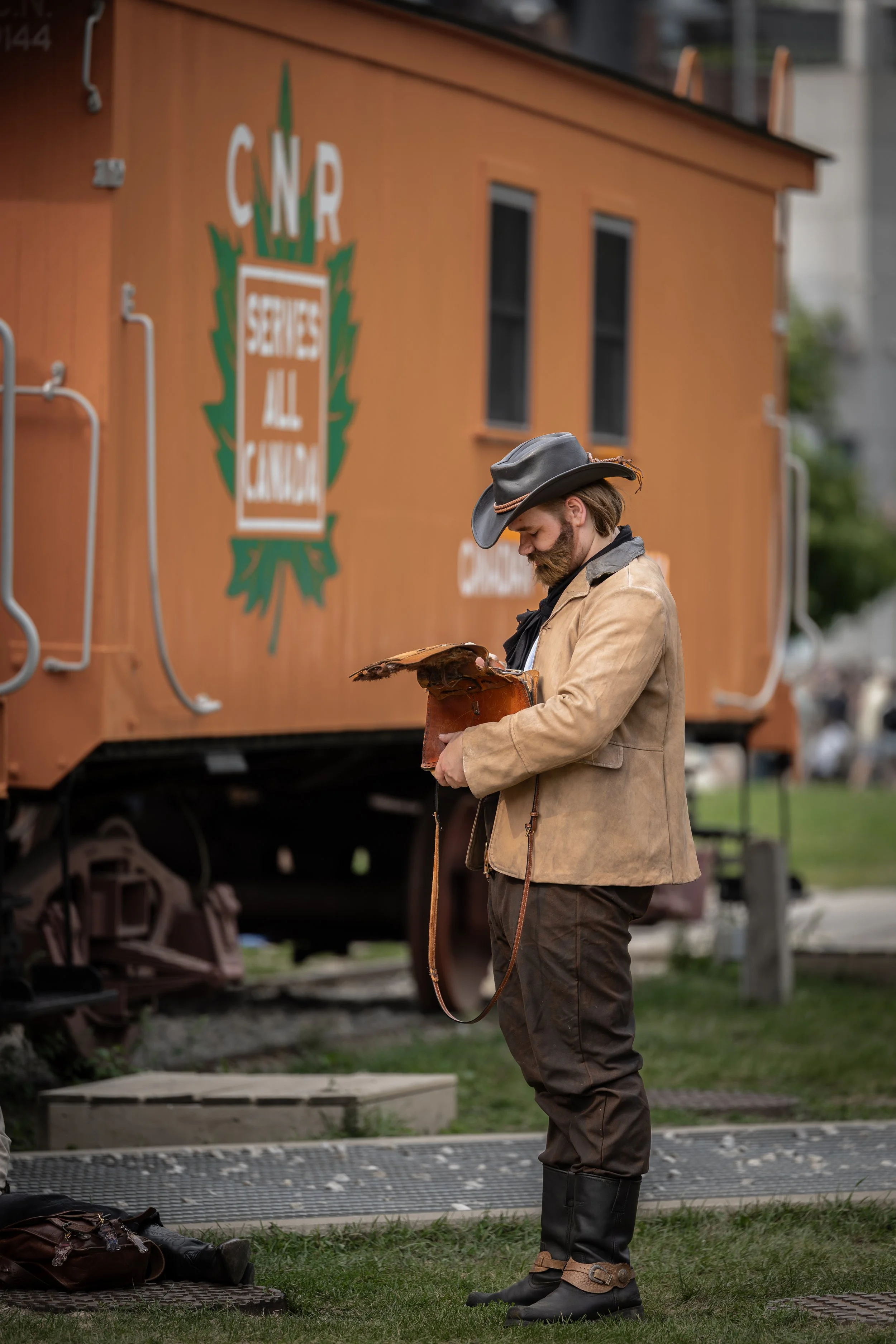 A man dressed in cowboy attire, including a leather jacket and cowboy boots, standing outdoors in front of a large orange train car with the sign 'CNR' and other text, holding a leather bag or pouch and reading or inspecting something.