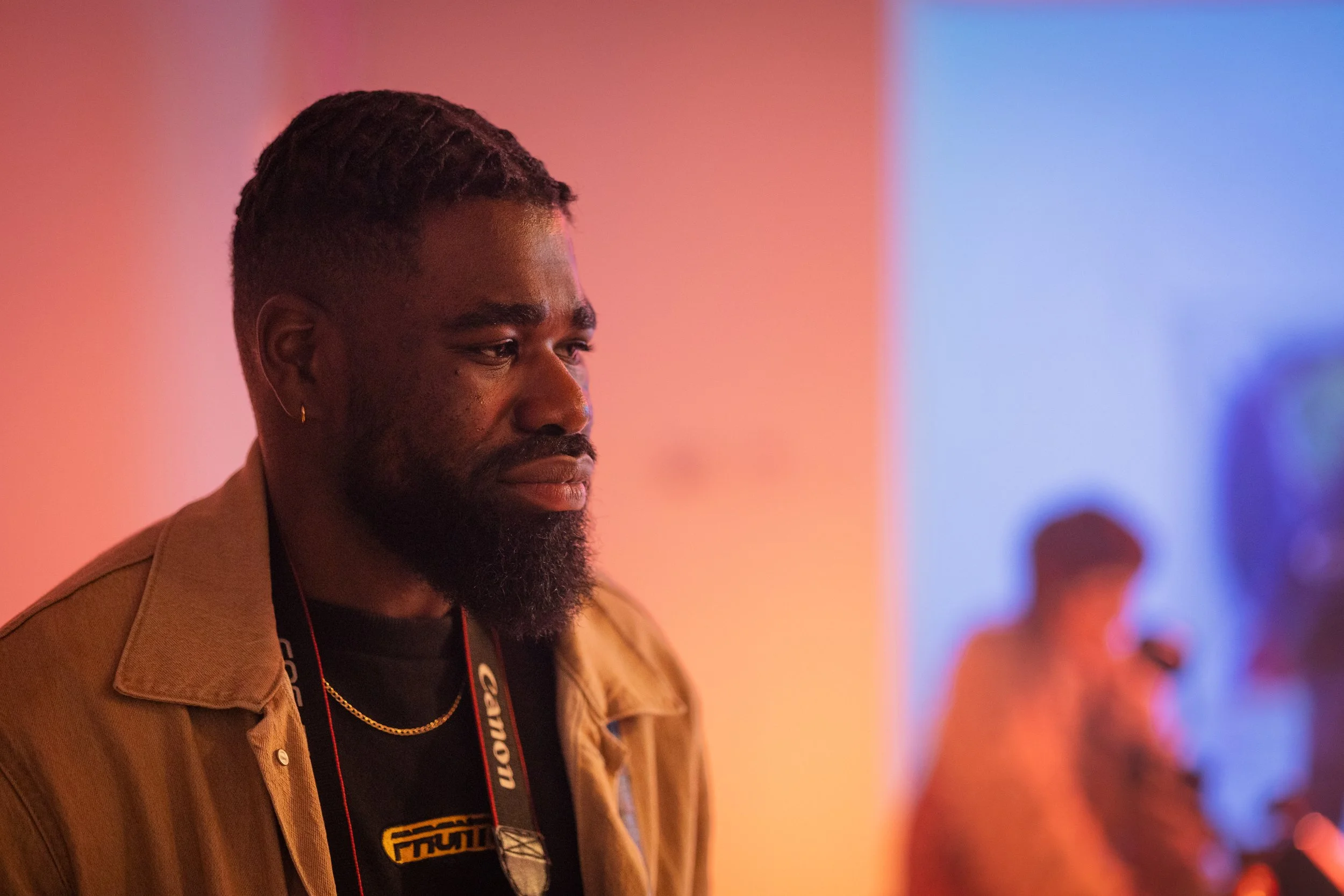 A young man with dark skin, a beard, and dreadlocks, wearing a tan jacket and a black T-shirt, looking down in front of a colorful blurred background.