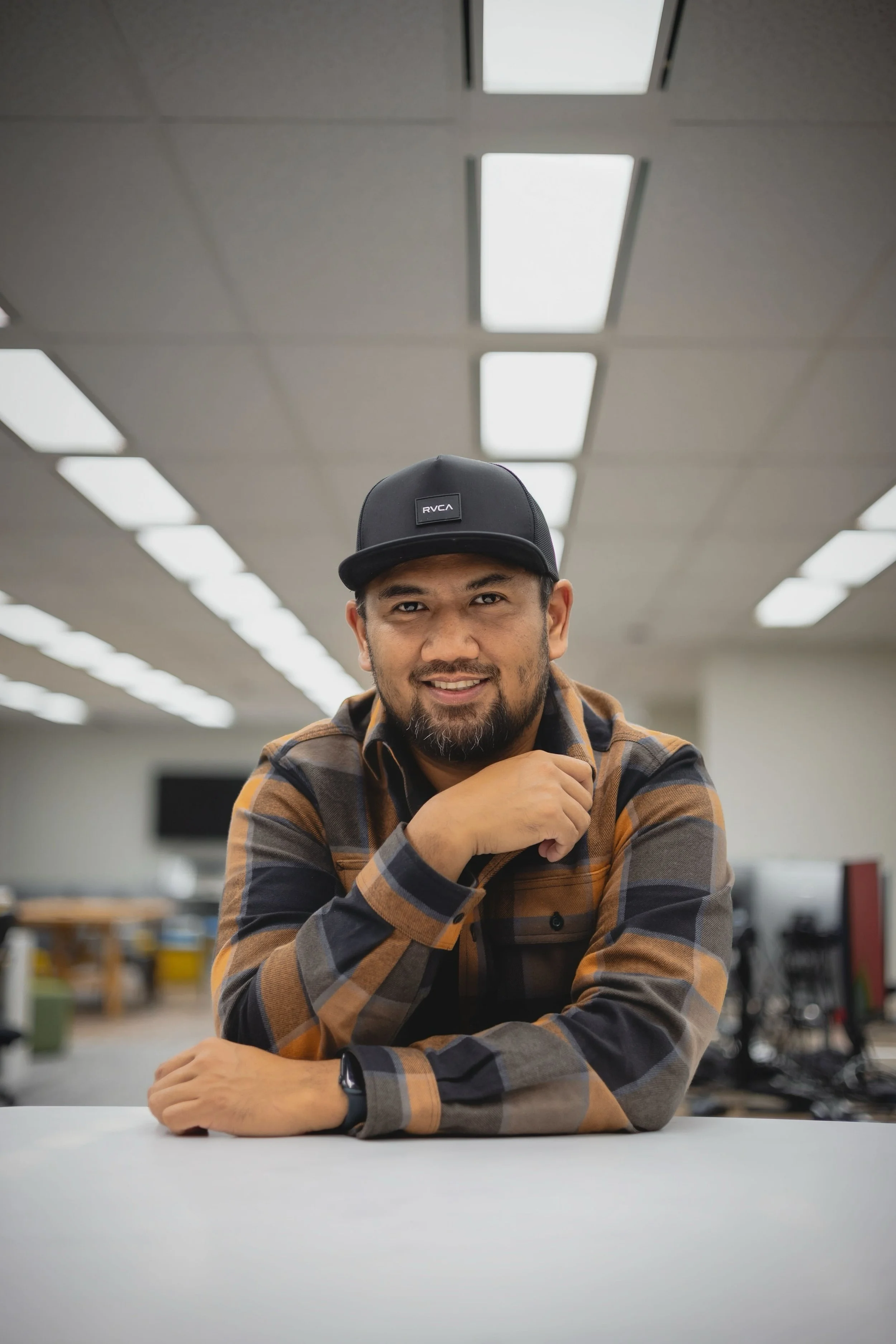 A man wearing a black RVCA cap, a plaid shirt, and a smartwatch, sitting at a white table in an office or classroom with a ceiling of fluorescent lights and blurred background.