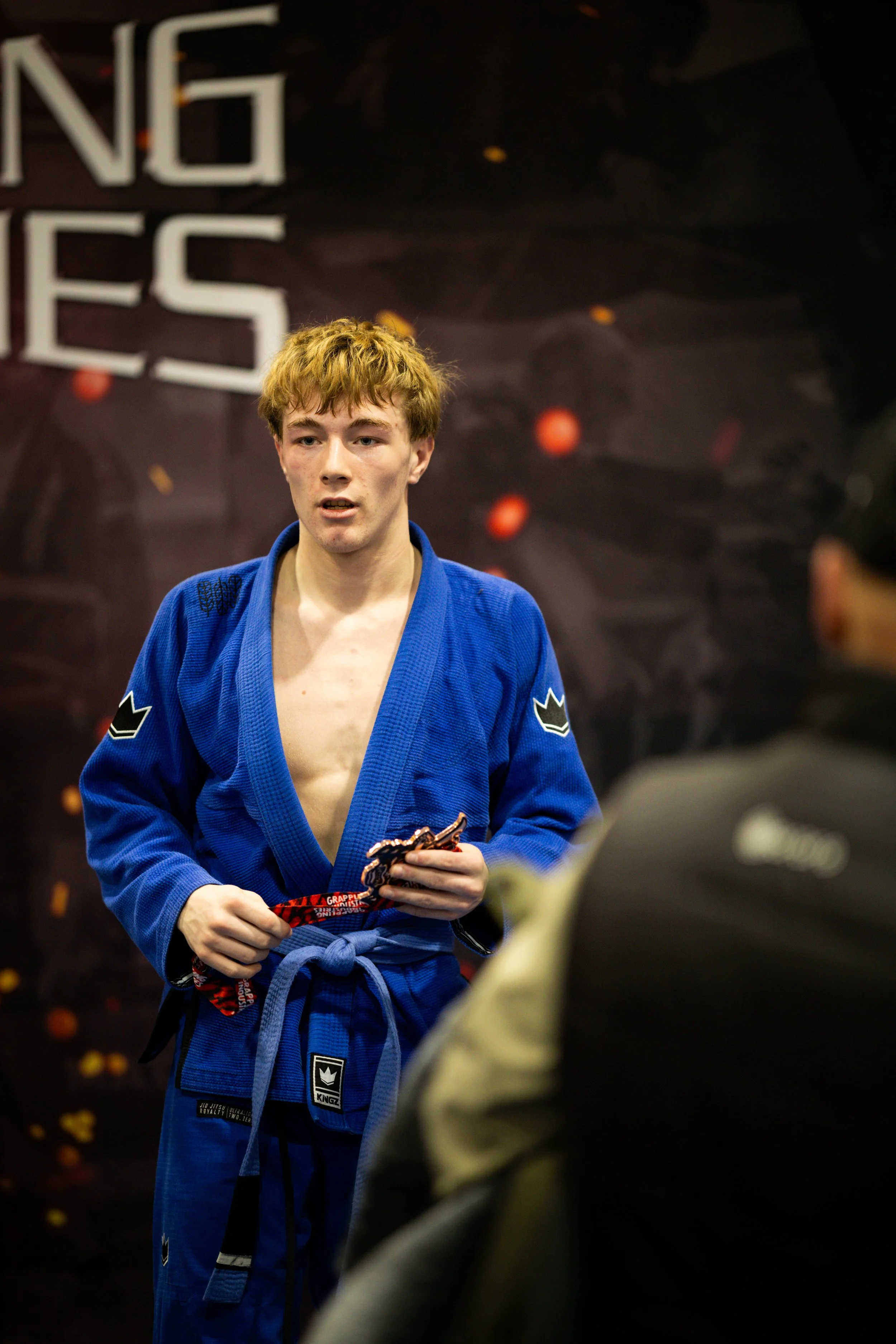 A young man with red hair and a focused expression wearing a blue Brazilian jiu-jitsu gi holding a red and black belt in a martial arts training facility.