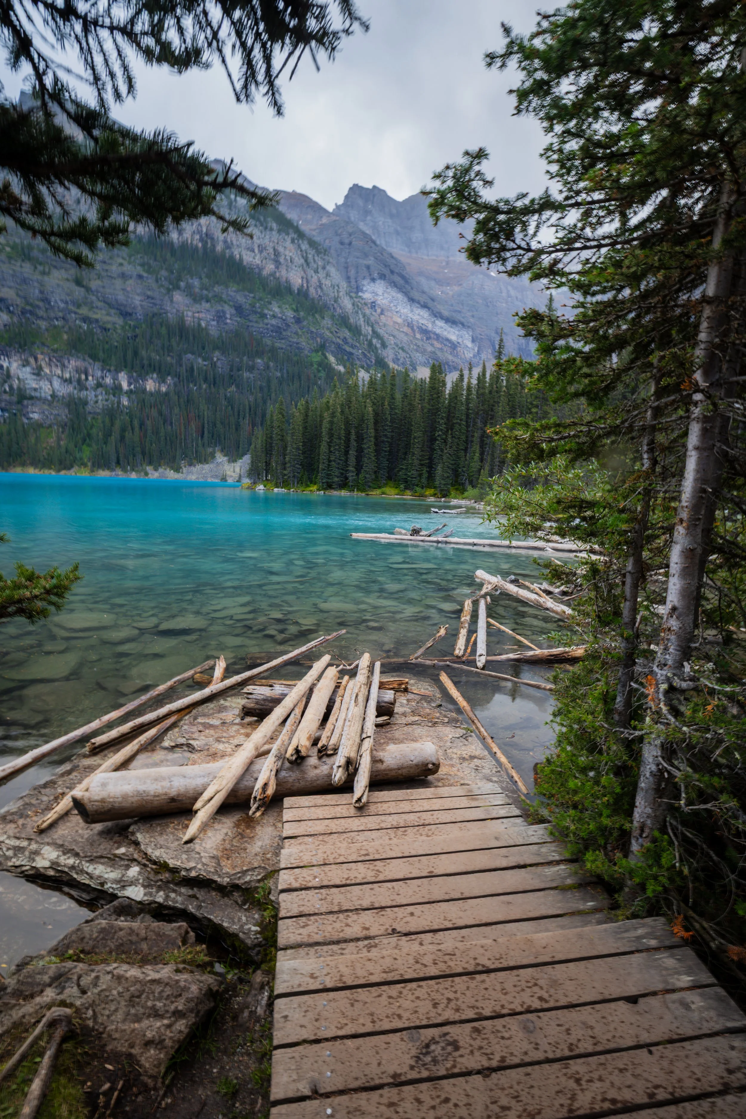 Wooden pathway along a rocky shoreline leading into a forest near a turquoise lake, with mountains in the background and trees framing the scene