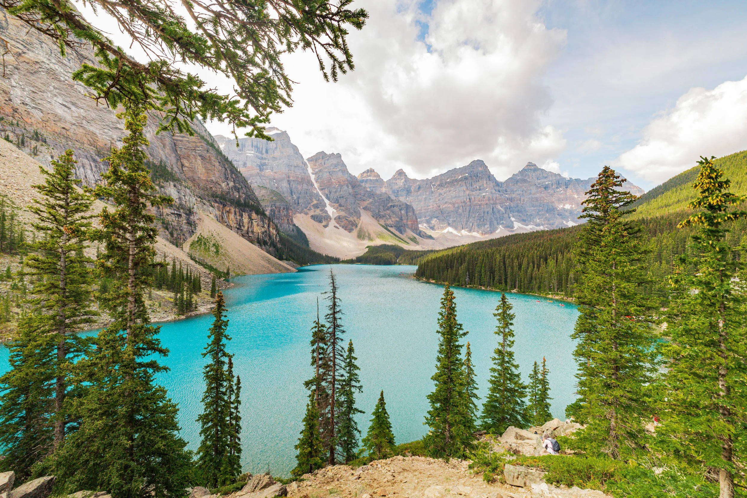 Scenic view of a turquoise lake surrounded by pine trees and rugged mountains with some snow patches, under a partly cloudy sky.