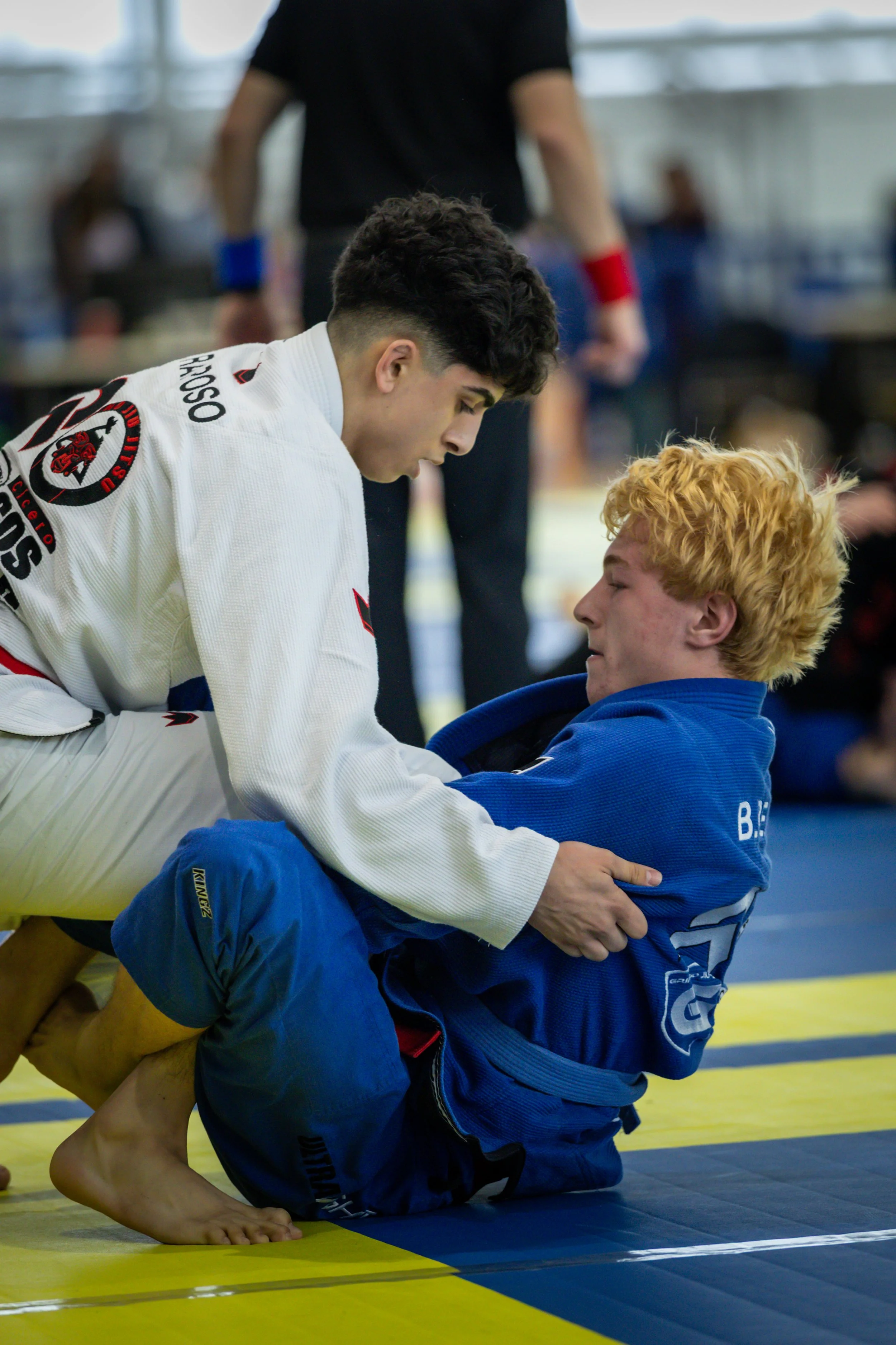 Two male Brazilian Jiu-Jitsu practitioners, one in a white gi and the other in a blue gi, engaged in a grappling move on a yellow and blue mat during a competition or training session.