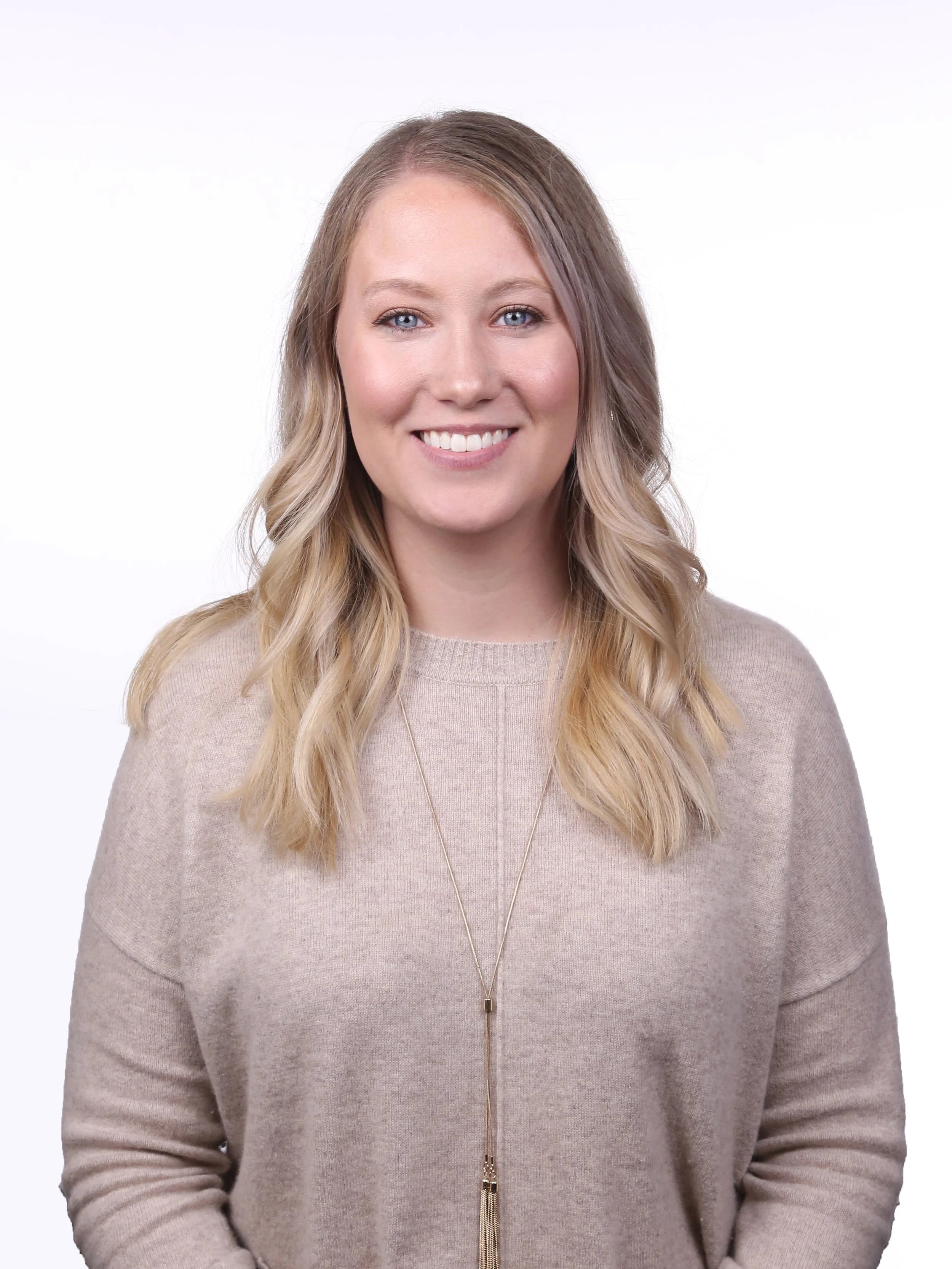 A woman with long wavy blonde hair, blue eyes, and light skin, smiling, wearing a beige sweater and a long necklace, against a plain white background.