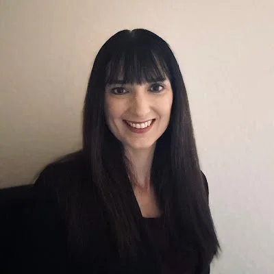 A young woman with long dark hair and bangs smiling at the camera, wearing a black top against a plain light-colored background.