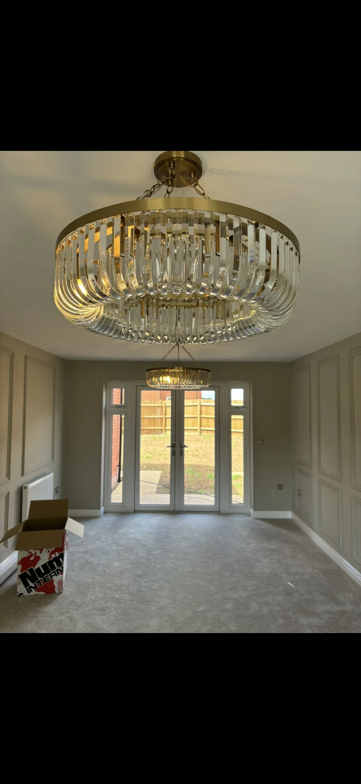 Modern chandelier with glass and gold accents hanging from a ceiling in a bright room with glass double doors leading outside, partially furnished with a cardboard box on the floor.
