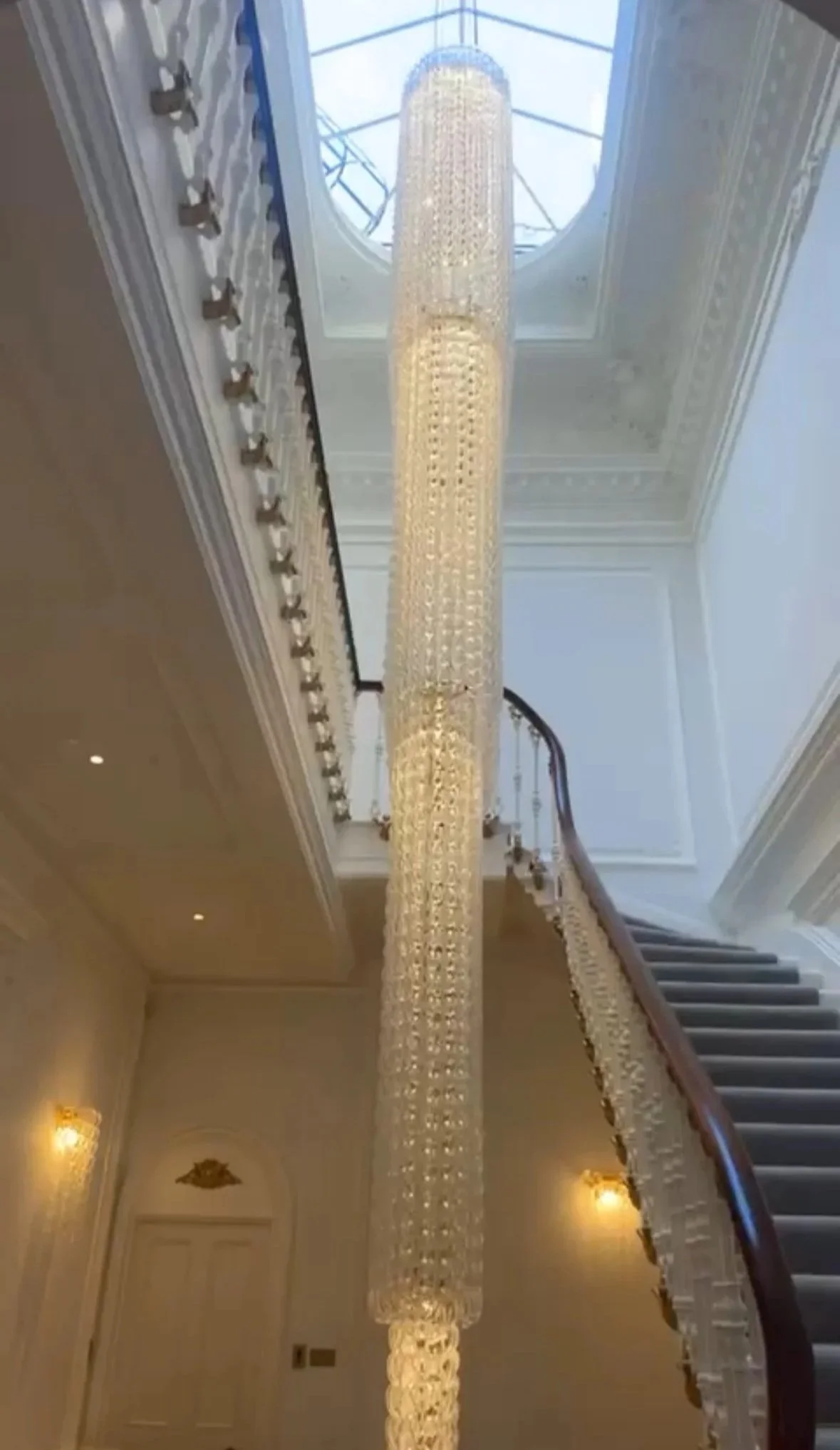 Large crystal chandelier hanging from the ceiling in a grand staircase area with white walls and a skylight above.