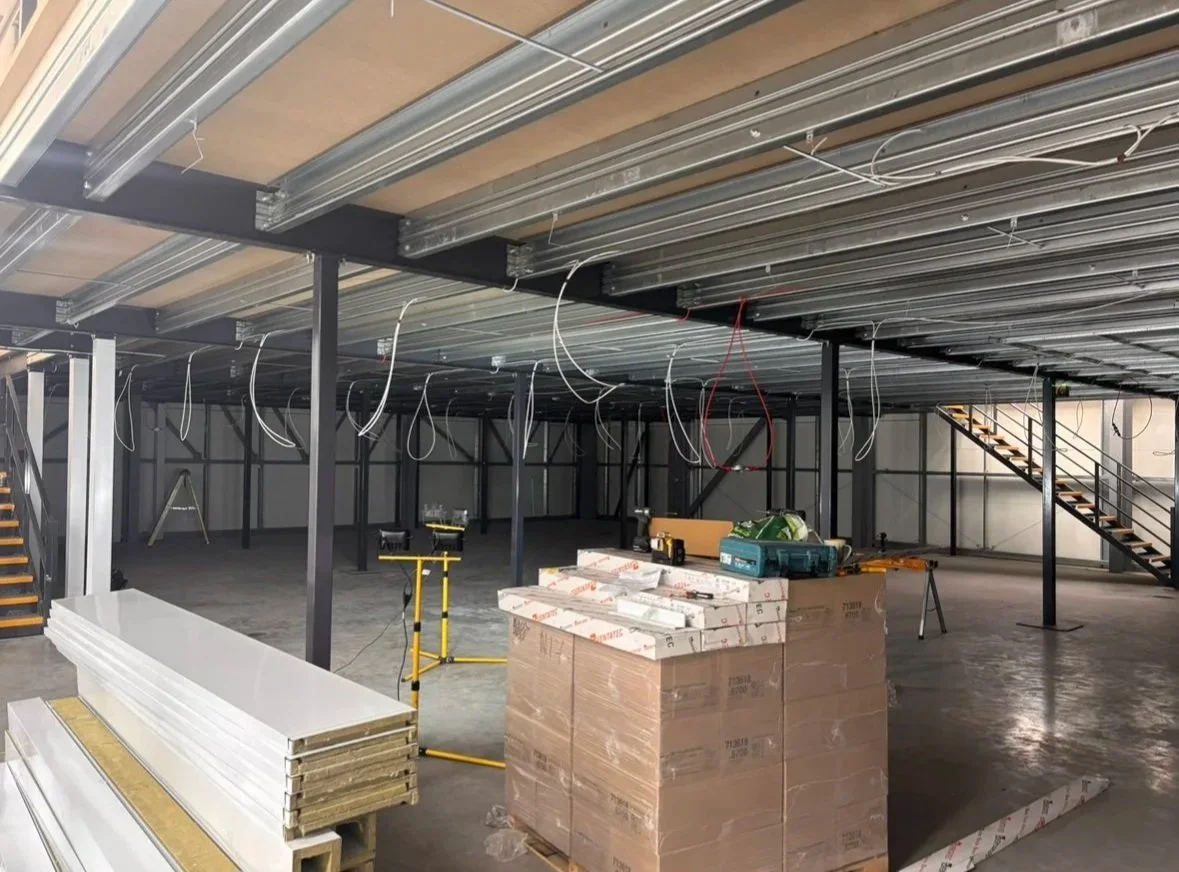Construction site with steel beams, staircases, and building materials, with electrical wiring hanging from the ceiling.