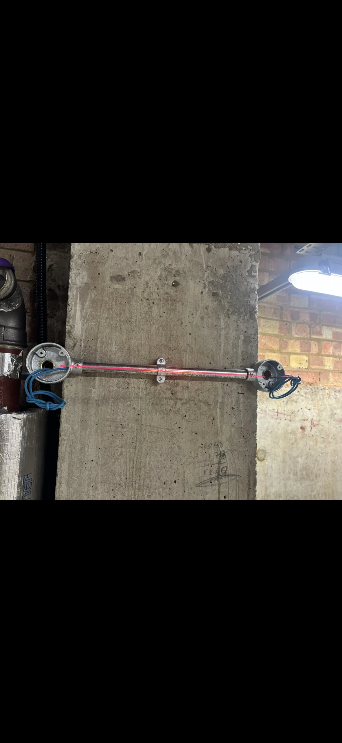 A laser device mounted on a wall with a blue wire connected to a circular component, with a concrete wall in the background and a bright light above.