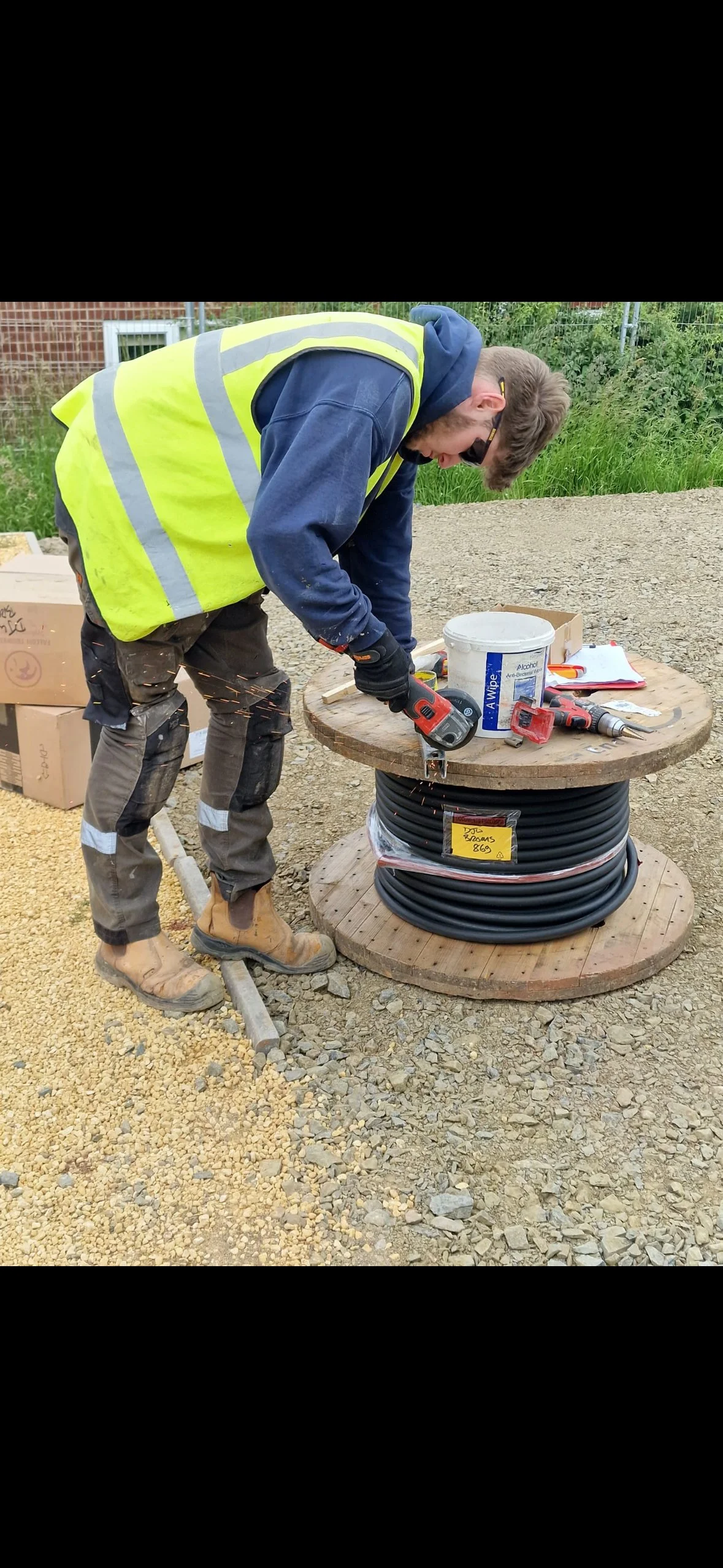 A construction worker in a high-visibility vest and work boots is cutting a large coil of cable on a wooden spool at an outdoor construction site.