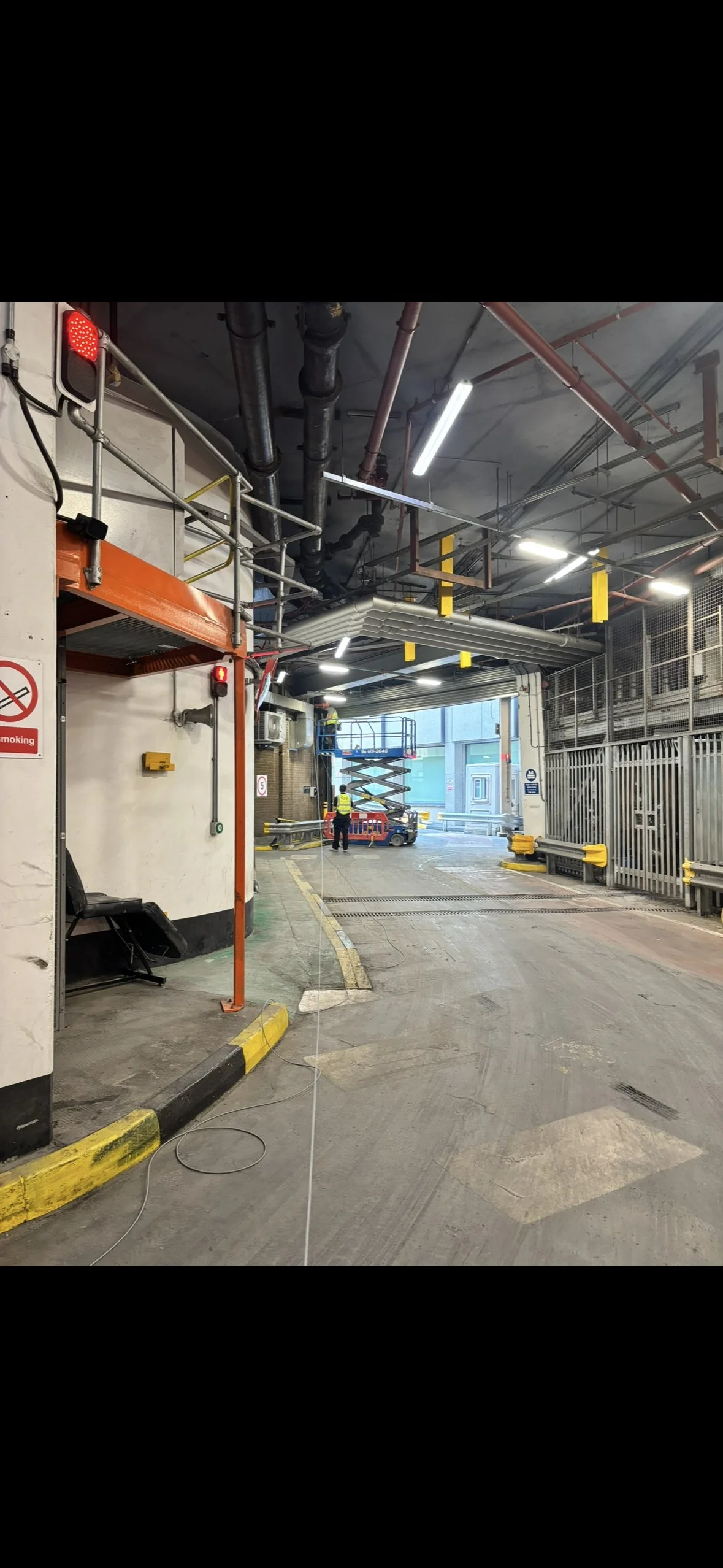 Indoor parking garage or loading dock area with a scissor lift, safety yellow barriers, and two workers in yellow vests. Open garage door revealing outside buildings.