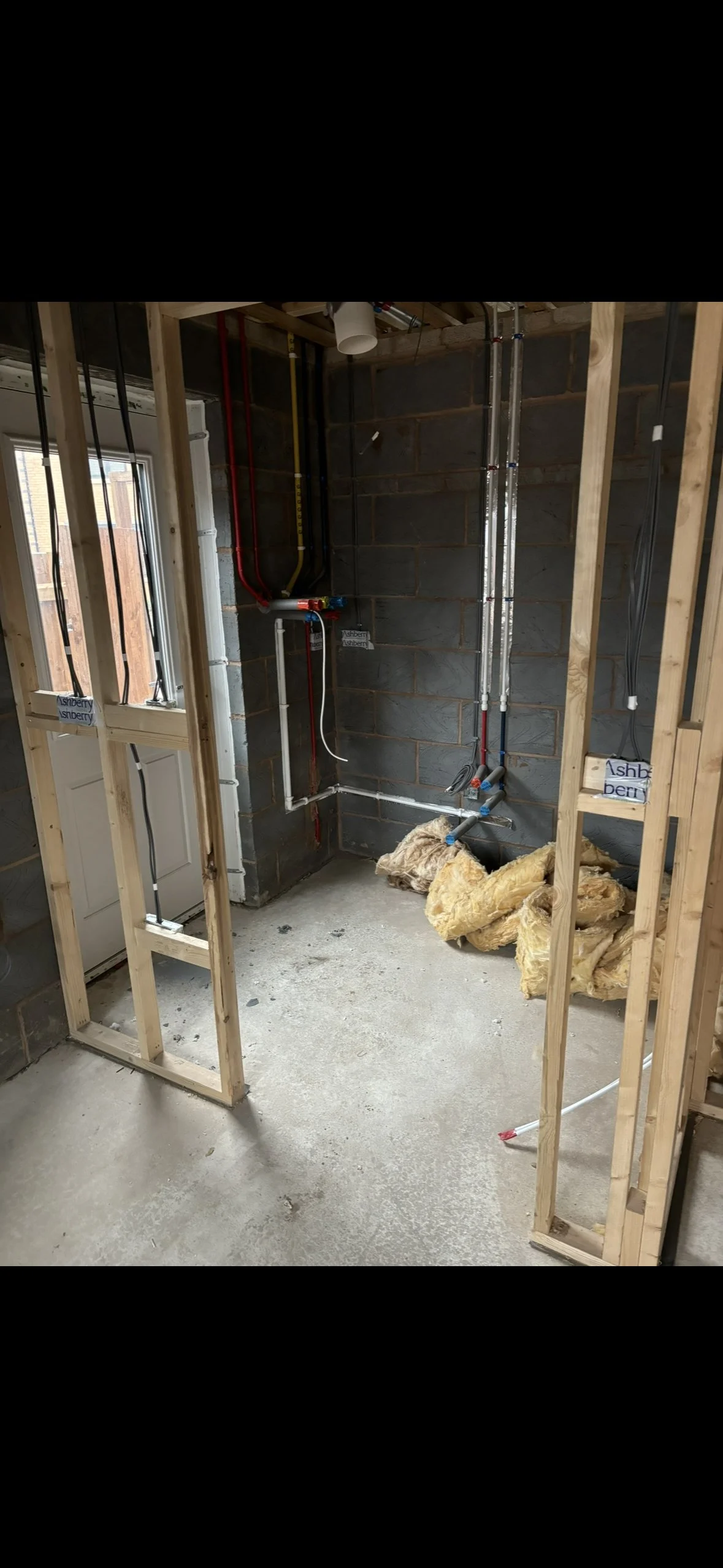 Interior of a room under construction with exposed wood framing, plumbing pipes, insulation on the floor, and a partially finished window.