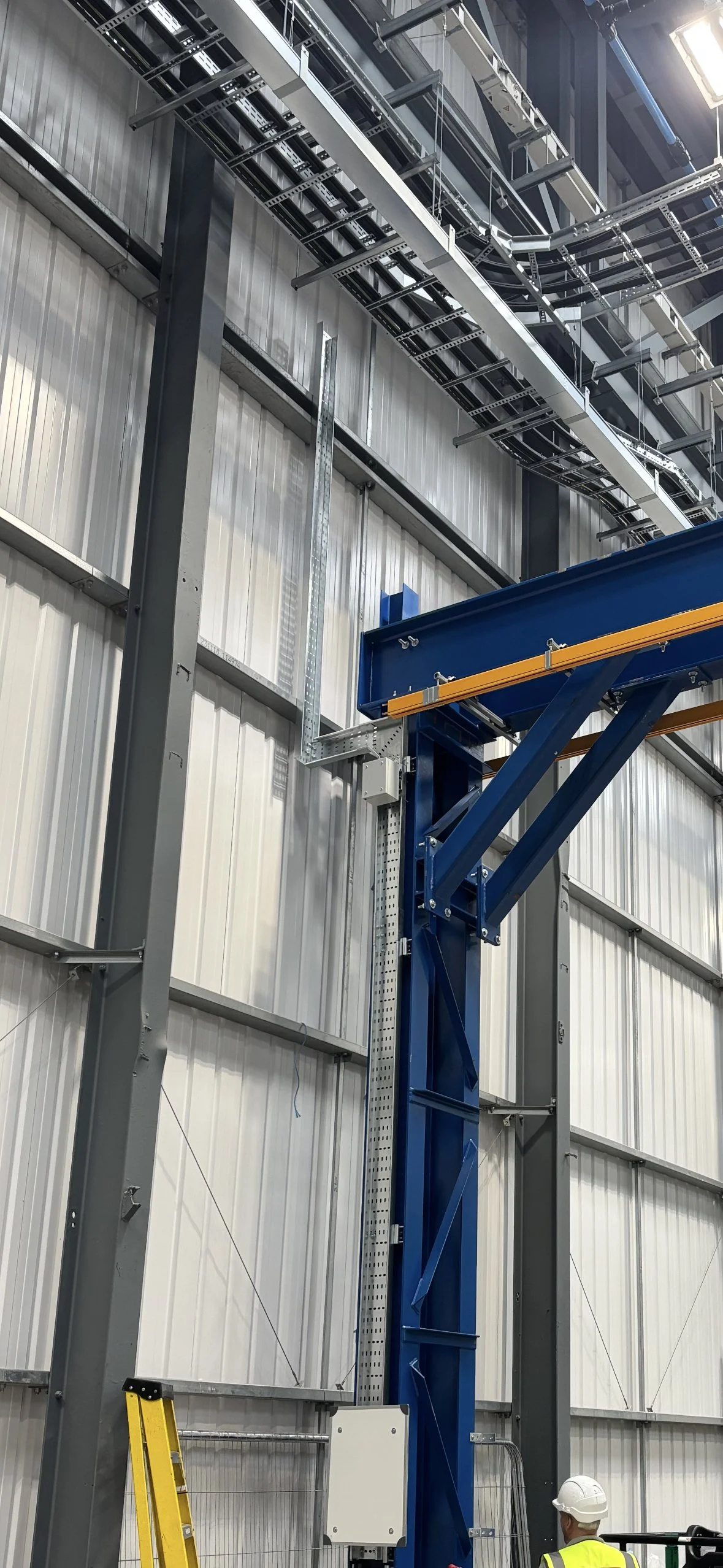 Inside an industrial warehouse, a construction worker wearing a white helmet and yellow safety vest stands near large blue and gray metal structural supports, with overhead cables and metal framework in the background.