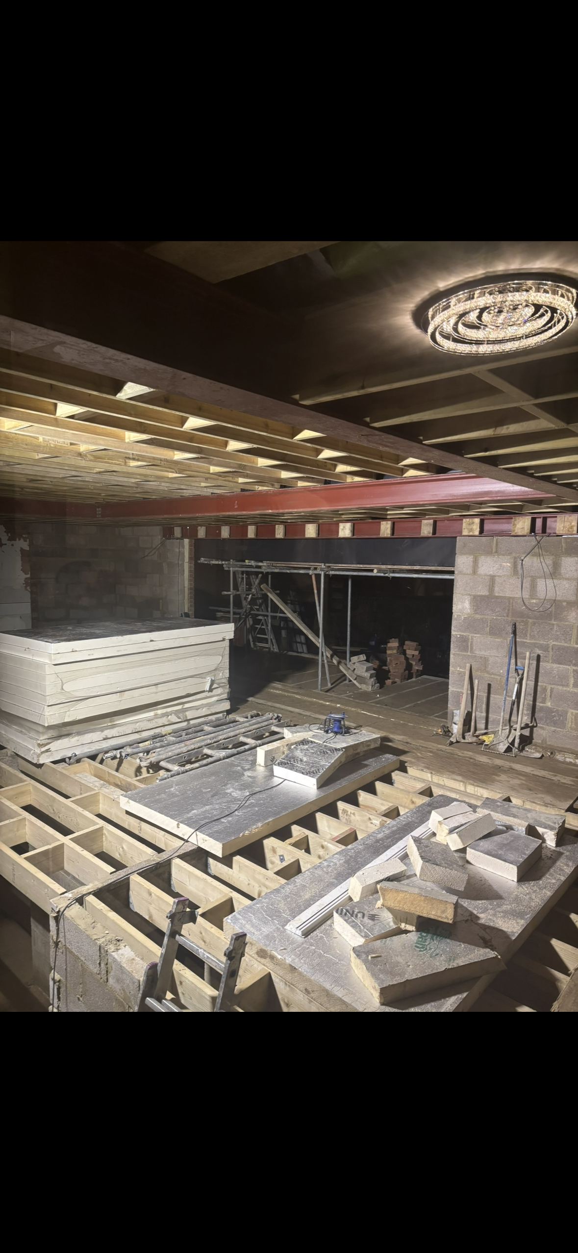Interior construction site with wooden framework for a floor or ceiling, construction materials, tools, scaffolding, and a decorative ceiling light.