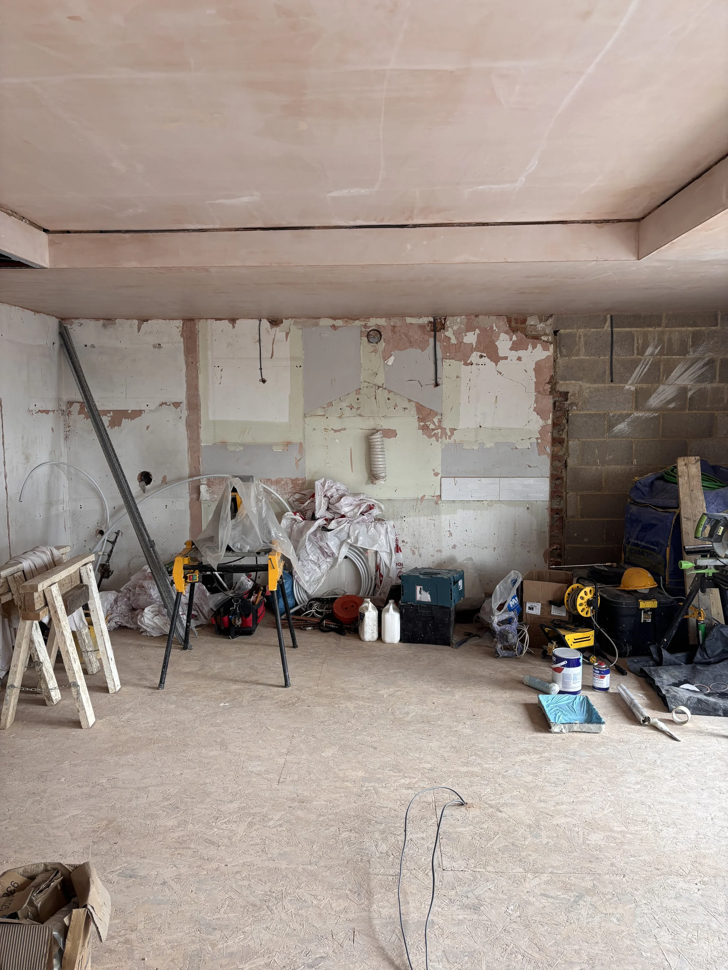 Construction site with tools, equipment, and materials scattered across the floor; unfinished walls and a large gap in the ceiling.