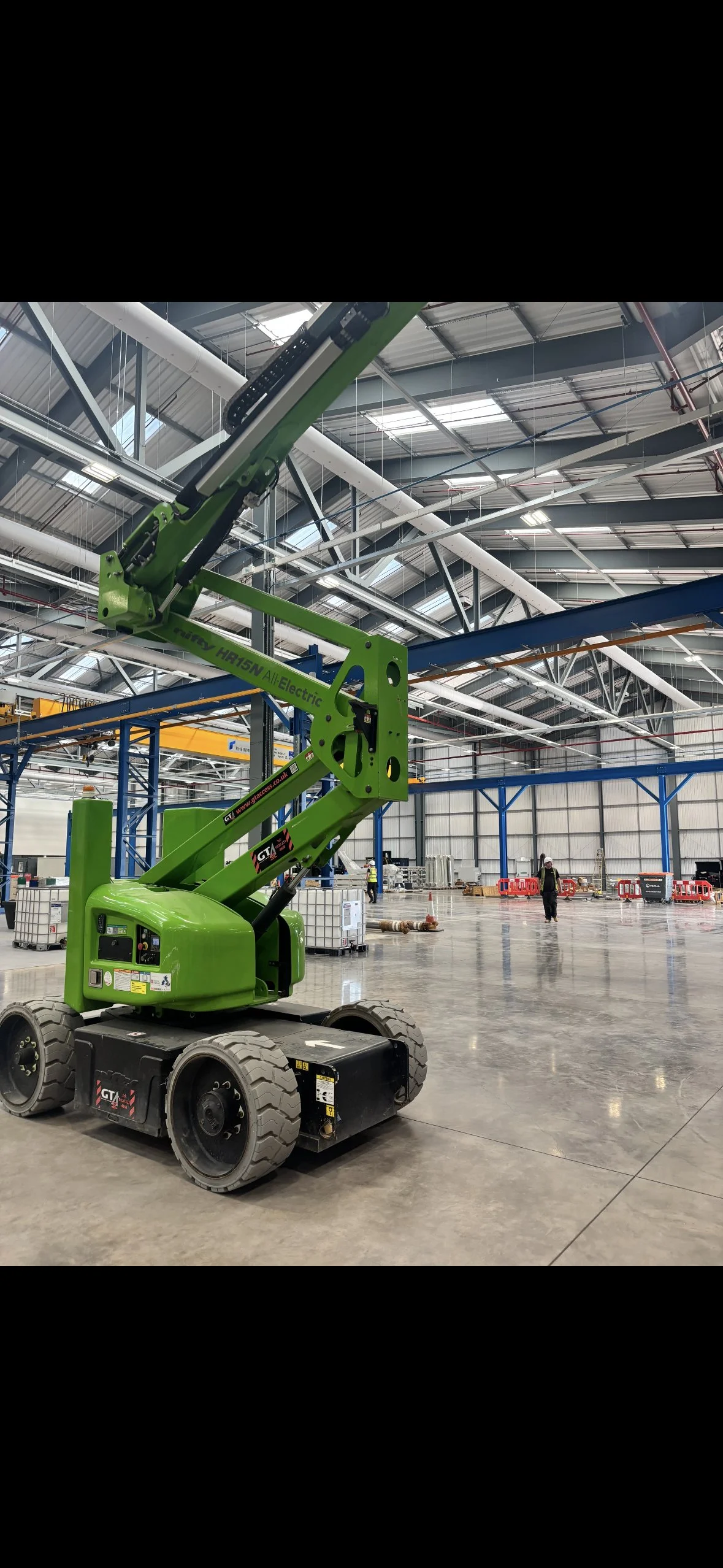 A green electric boom lift inside a large industrial warehouse under construction, with workers and construction equipment in the background.