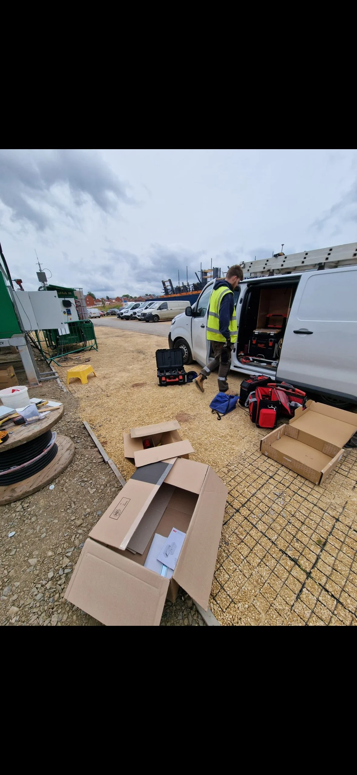 A man in a yellow safety vest working with tools in the open side of a white van, surrounded by toolboxes, boxes, and equipment on the ground, with parked cars and industrial equipment in the background.