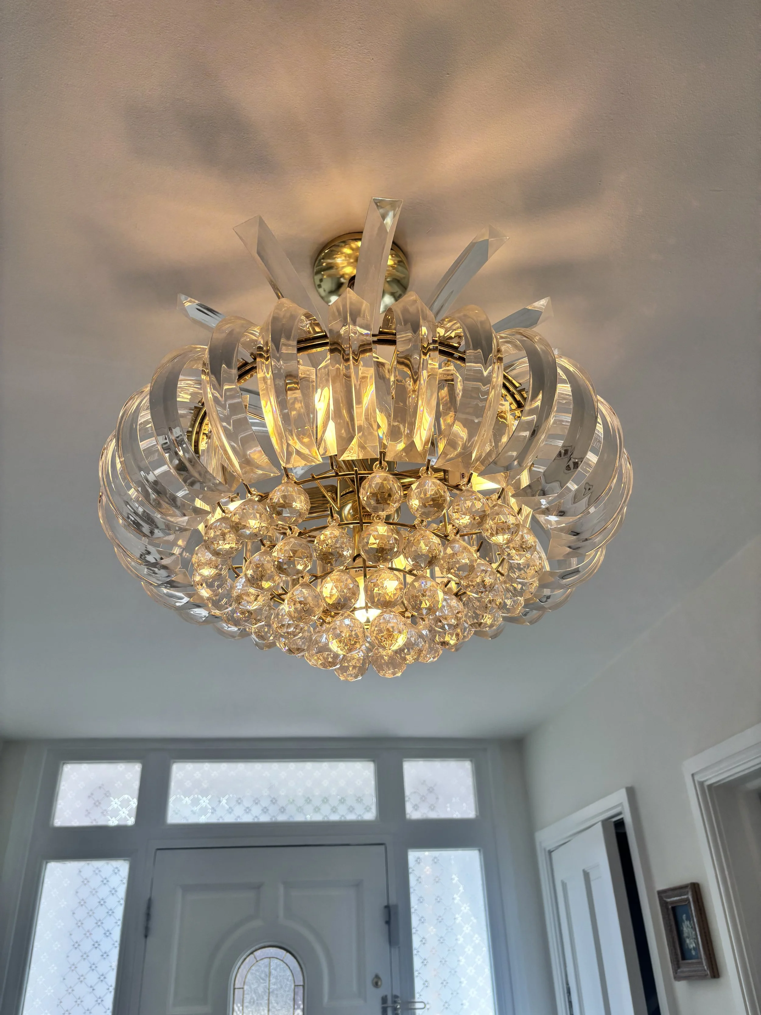 Chandelier with crystal and glass ornaments hanging from a ceiling, illuminating a white entryway with a front door and large side windows.