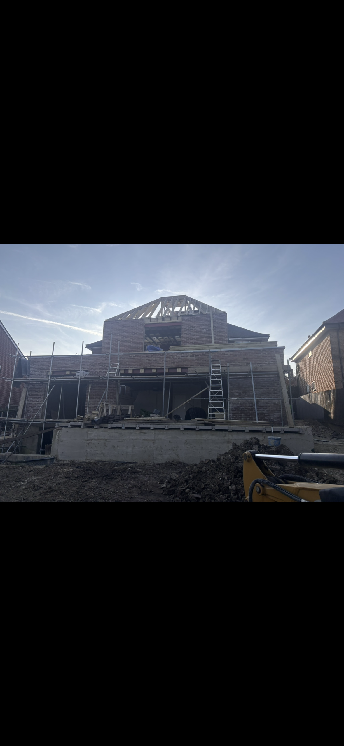Construction site of a house with brick walls and a wooden roof framework, scaffolding surrounds the building, and construction materials are on the ground.