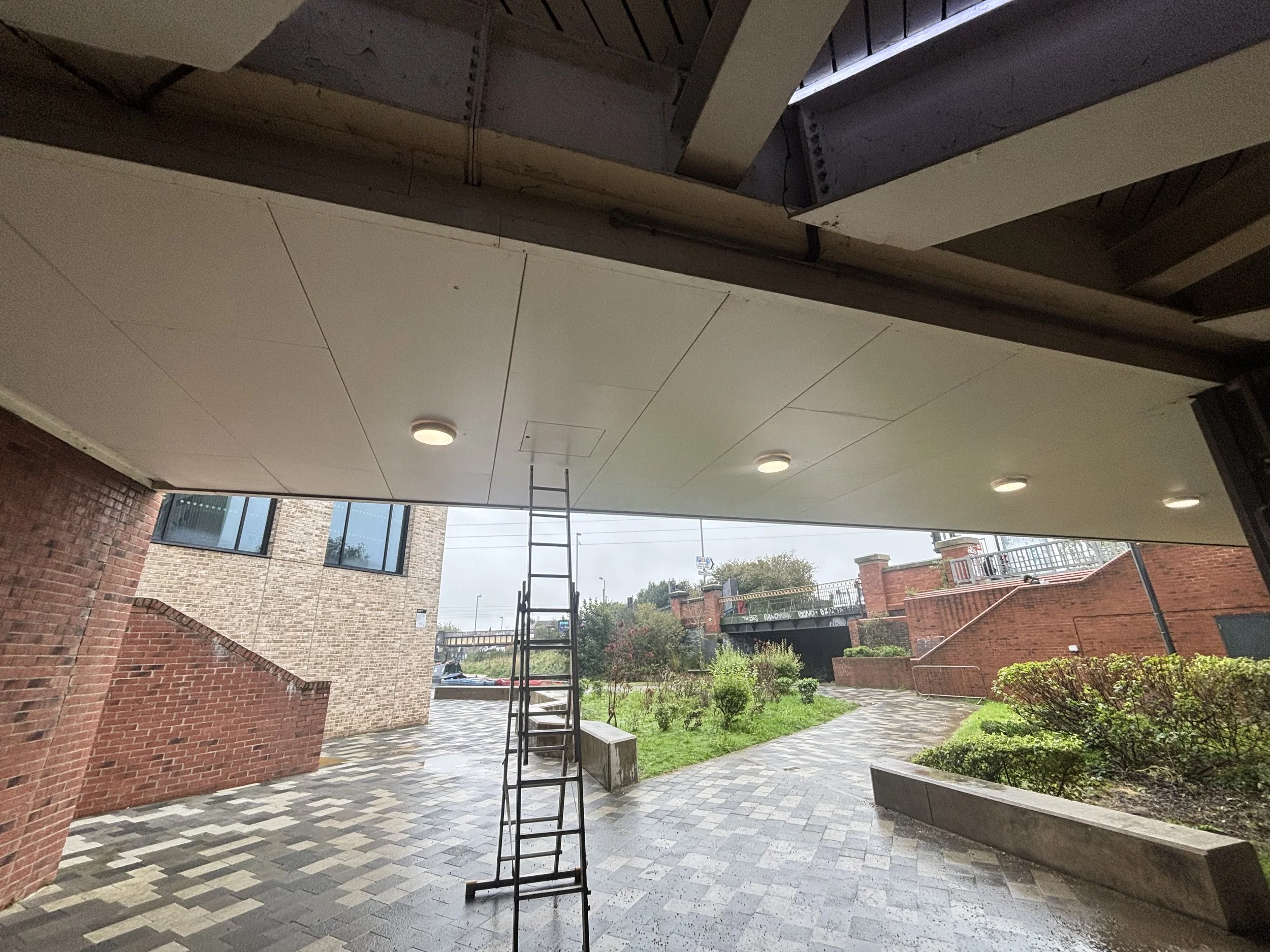 Underneath a covered outdoor patio with a tiled floor, a step ladder, brick wall, and view of a garden and street outside.